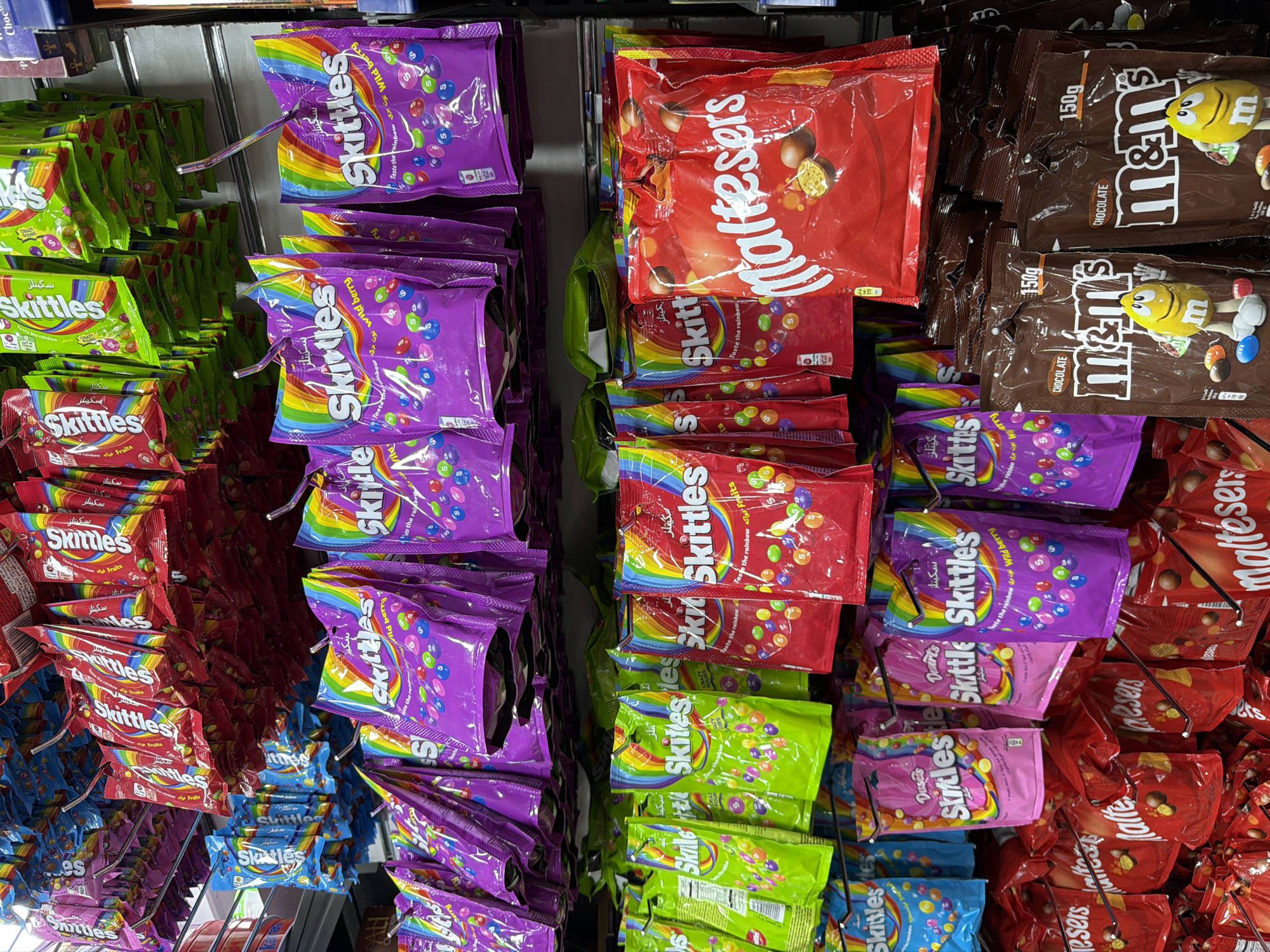 The image depicts a retail display of various candy products, predominantly Skittles and M&M's, on shelves in a store. The candies are packaged in colorful bags and pouches, with different flavors and varieties visible. Some bags of Skittles feature a rainbow design and the text "Taste the rainbow." M&M's bags are brown with the M&M's logo and cartoon characters. The store appears to be well-stocked, with multiple rows of candy products. The lighting suggests an indoor environment. The location is identified as Sharm El Sheikh, Egypt.