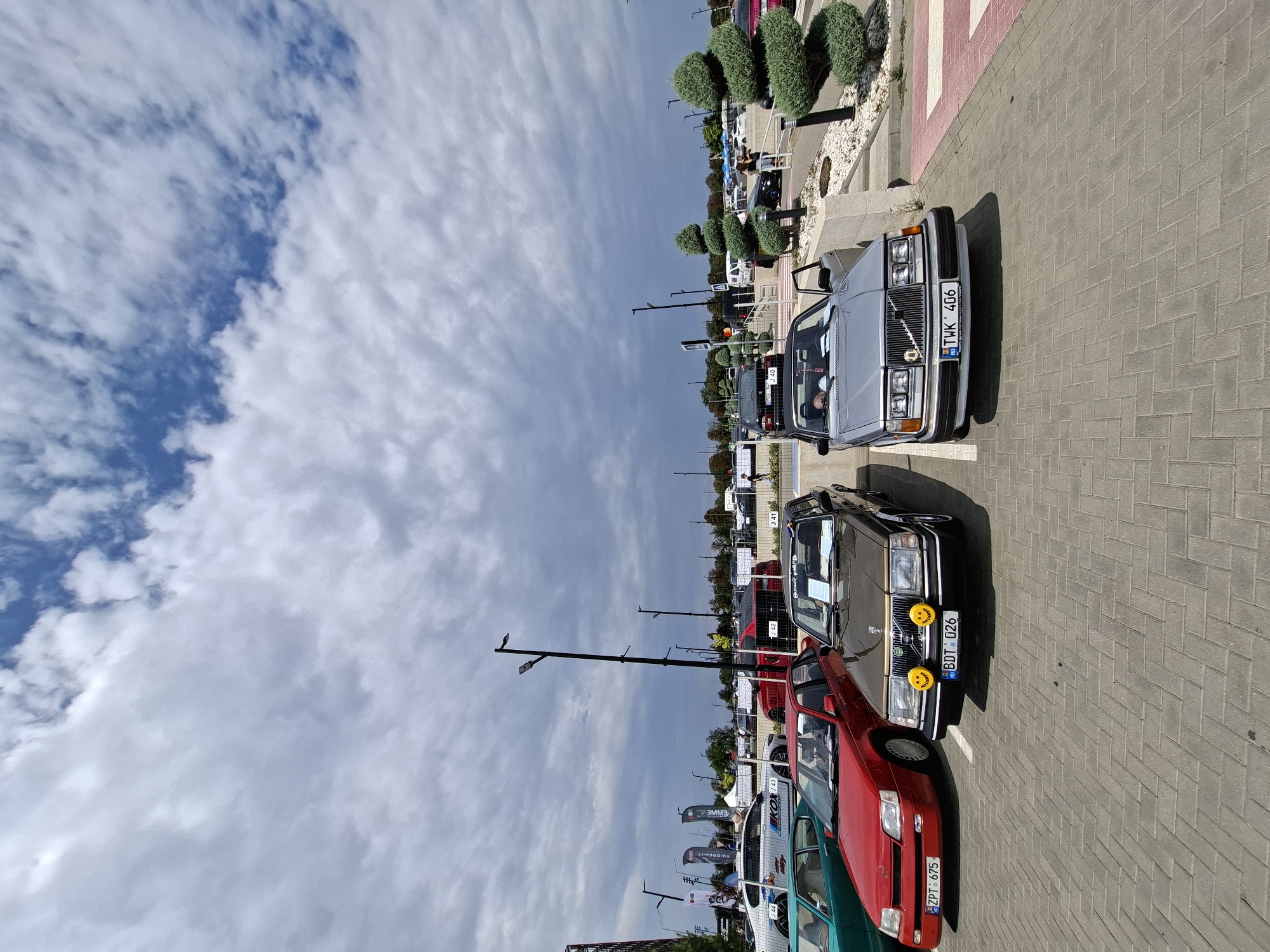 The image shows an outdoor car exhibition with several vintage cars parked on a paved surface. The sky is partly cloudy, with a mix of blue sky and grey clouds. In the foreground, three cars are prominently displayed: a red Opel, a brown Volvo, and a silver Volvo. The red Opel has a license plate that reads "ZPT 675". The brown Volvo has two yellow smiley face stickers on its grille and a license plate reading "BDT 026". The silver Volvo has a license plate "TWK 406". In the background, more cars are visible, as well as tents, flags, and people walking around. The exhibition appears to be taking place in a parking lot or a similar open space. The presence of various car models suggests a car show or event. The location is identified as Stăuceni, Moldova, likely an outdoor venue hosting this car event.