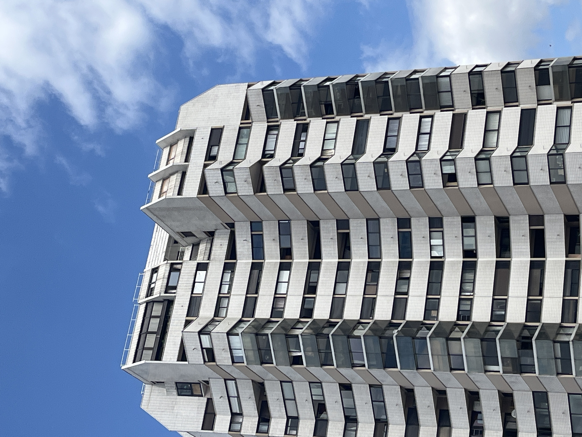 The image shows the exterior of a tall, modern apartment building in the city of Créteil, France. The building features a facade of white tiled panels and beige bands that create a staggered, geometric pattern. Numerous windows and balconies are visible on each level, indicating a residential structure. The building is set against a bright blue sky with scattered white clouds. The perspective is from below, looking up at the building. The scene is devoid of people or any discernible activity.