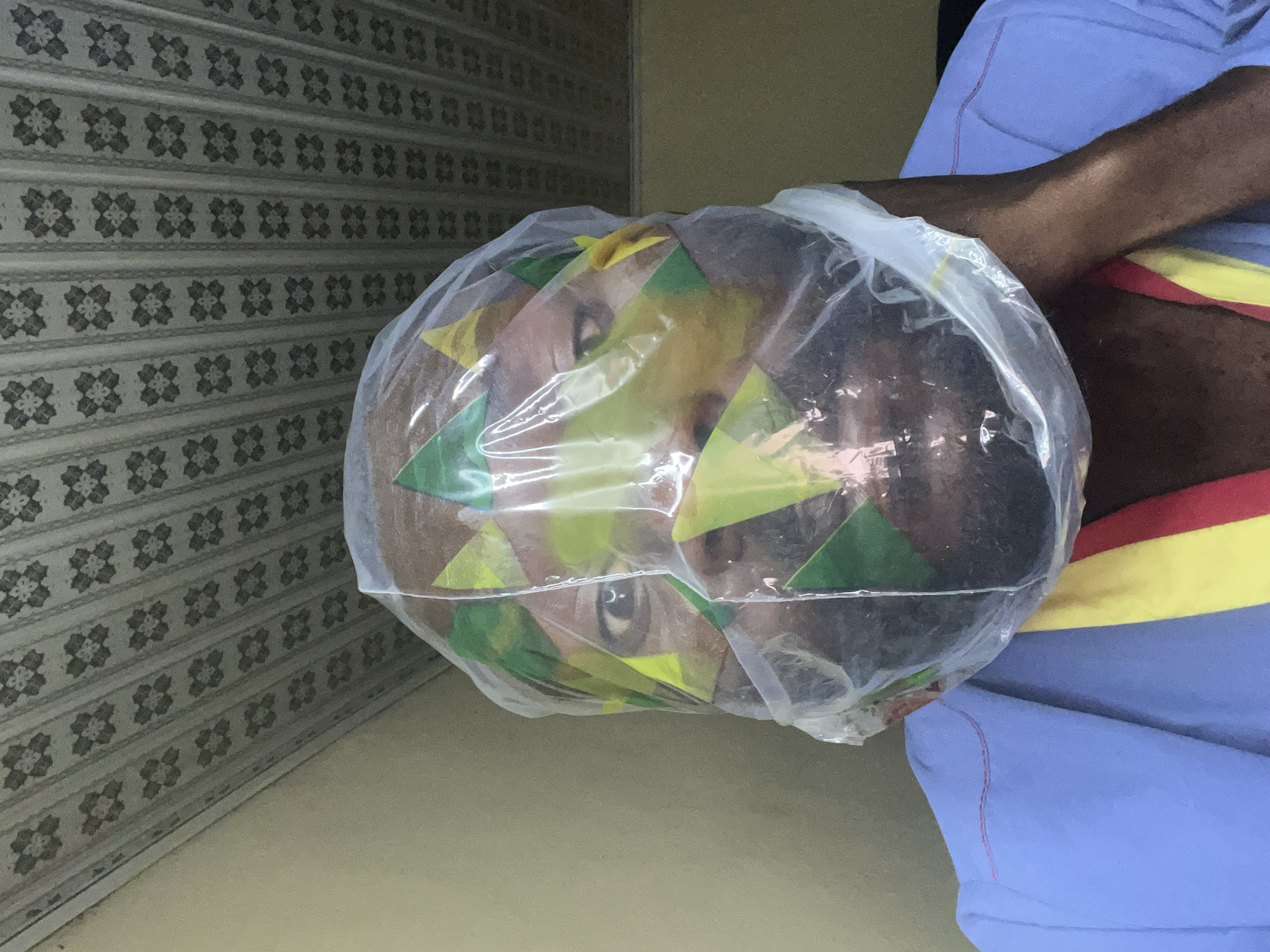 The image captures a close-up indoor scene featuring a person whose head is encased within a clear plastic bag. Underneath the transparent bag, various geometric shapes in vibrant green and yellow colors, possibly paper or fabric, are visible around the person's head. One eye is distinctly visible through the plastic, looking forward. The person has dark skin.
To the right, a dark-skinned arm is visible, partially clad in a blue garment that features horizontal stripes in red and yellow near the torso. The background consists of two distinct sections. On the left, a wall or shutter features a repeating pattern of dark gray, stylized floral or geometric motifs against a lighter, possibly off-white or beige, background. On the upper right, a plain light tan or beige wall section is visible.
There is no specific activity or event clearly depicted, though the head covering suggests a potential costume, performance element, or a unique form of personal expression. No text is visible in the image. The setting appears to be indoors with artificial lighting. The location context provided is Gura Top, Nigeria.