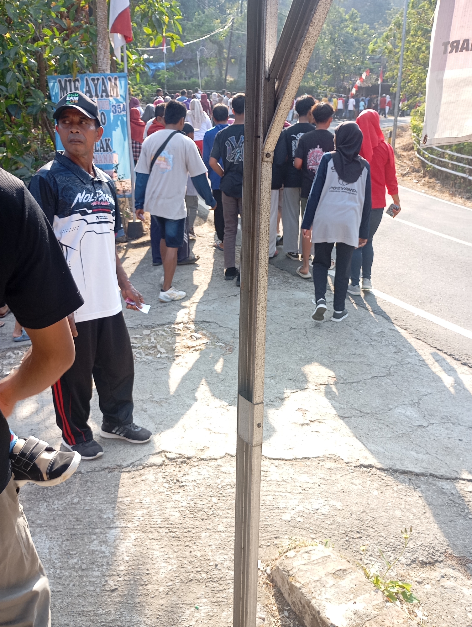 In Ketepung, Indonesia, a group of people are gathered on a paved walkway adjacent to a road. The scene is set outdoors, with trees and foliage visible in the background. A man in the foreground, wearing a black and white patterned shirt and a black cap, is looking to his left. He holds a small white object, possibly money, in his right hand. Behind him, several individuals are walking away from the camera, some in casual attire and others in athletic wear. A blue sign with white text, partially obscured by the man, reads "MIE AYAM" and other Indonesian text. A red and white flag is visible on a pole to the left of the sign. The road to the right is lined with a white stripe. The ground is cracked and worn, with patches of grass growing through. Sunlight casts shadows on the walkway, indicating it is daytime.