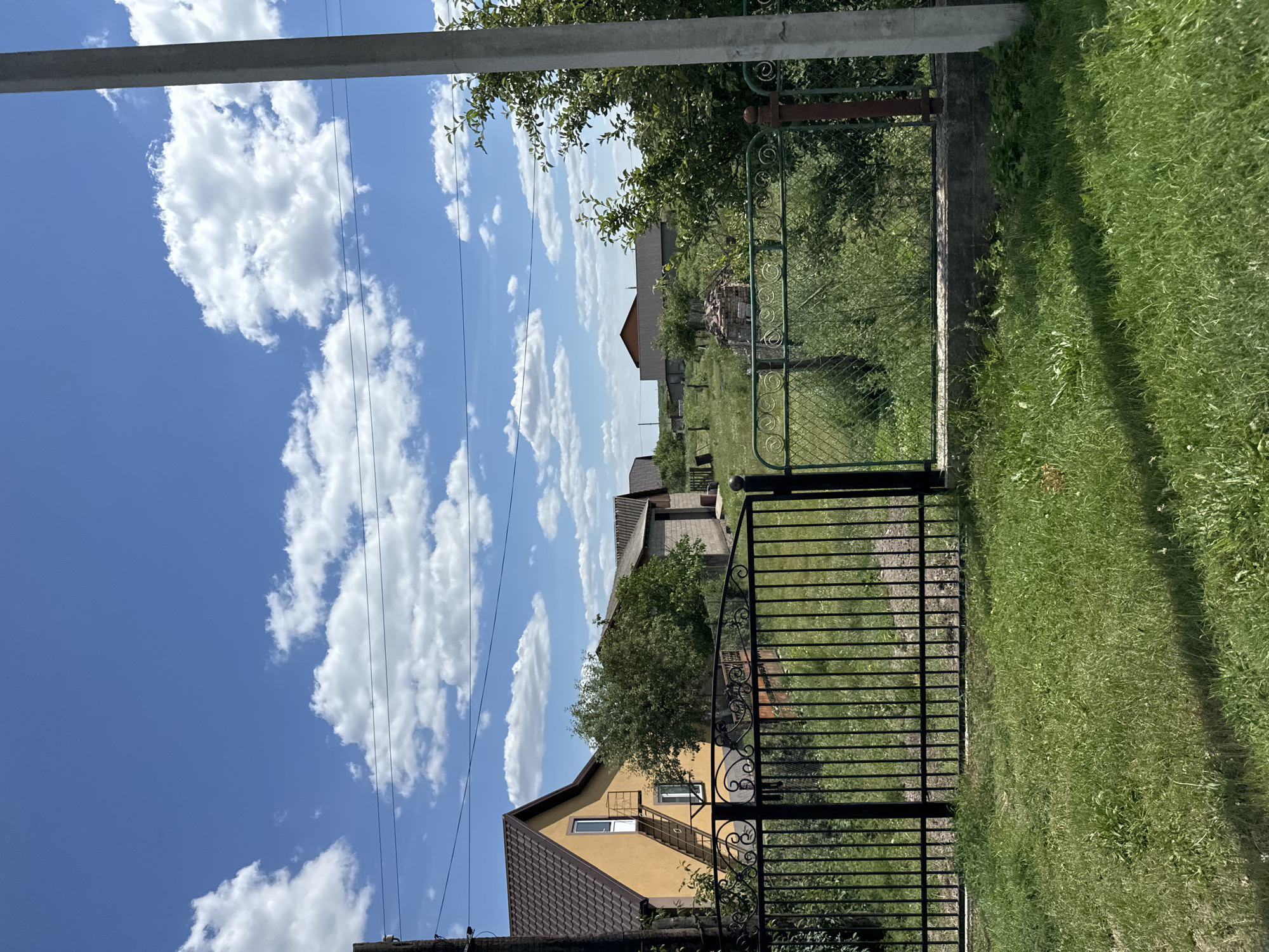 The image depicts a rural landscape under a bright, partly cloudy sky. In the foreground, a black wrought-iron fence stands partially open, revealing a grassy yard beyond. To the left, a yellow house with a dark brown roof and an external staircase is visible. Behind the fence, various structures are scattered across the property, including what appears to be a brick building and several other houses with tiled roofs in the distance. Lush green trees and vegetation are prevalent throughout the scene. A concrete utility pole stands prominently on the right side of the image, with wires running across the sky. The overall impression is that of a serene countryside setting, likely in the village of Dovhe, Ukraine, given the architectural style and the presence of utility poles and fencing common in such areas. No people or animals are present in the image.
