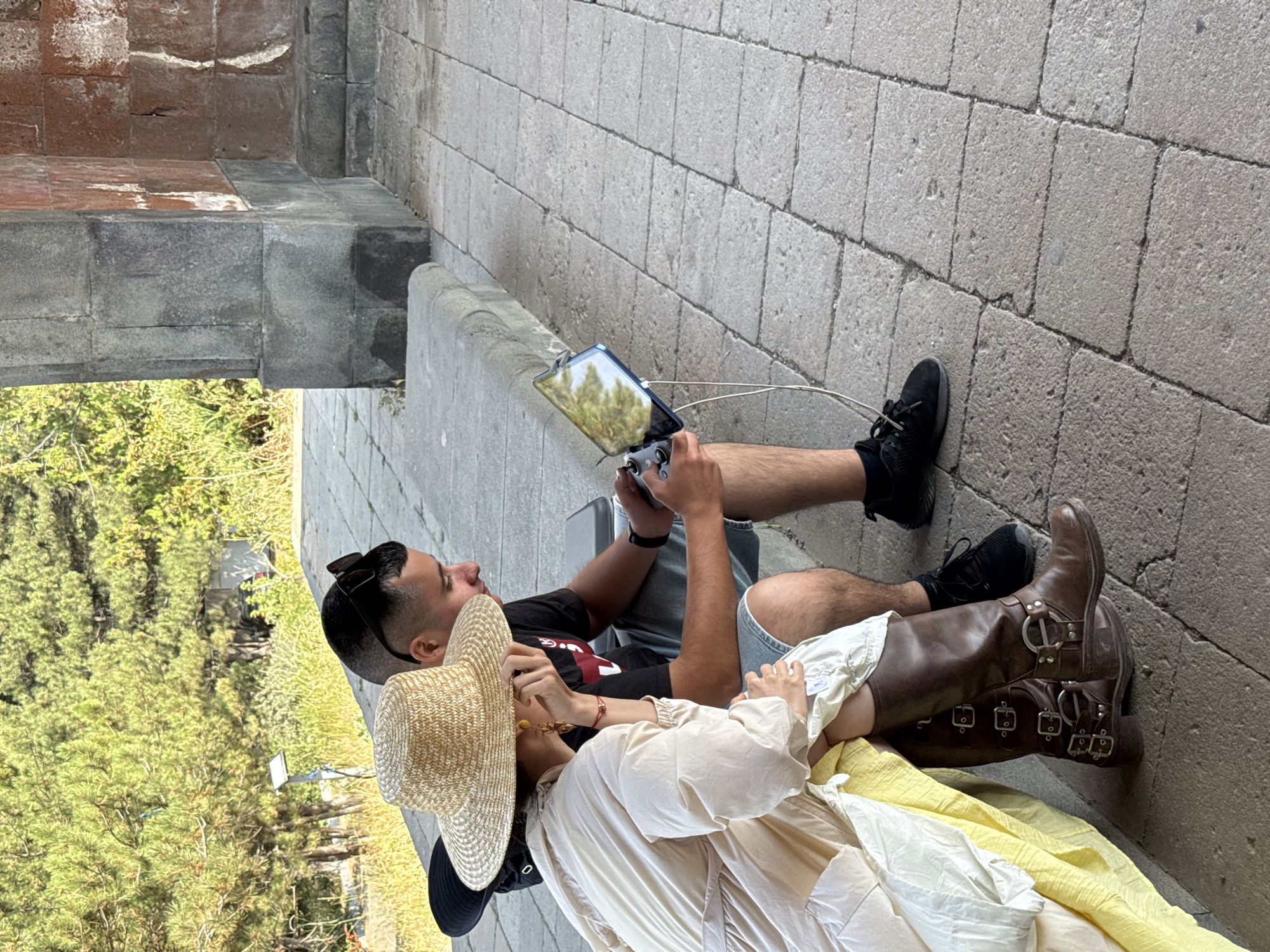 In Voghjaberd, Armenia, two individuals are seated on a stone ledge outdoors. A person on the left, wearing a large straw hat and a flowing light-colored dress with a bright yellow underskirt, is looking down, resting their chin on their hand. To their right, a person in a black t-shirt and denim shorts is holding a tablet connected to a drone controller and appears to be looking at the tablet. The surrounding environment consists of a stone-paved area and a backdrop of green trees.