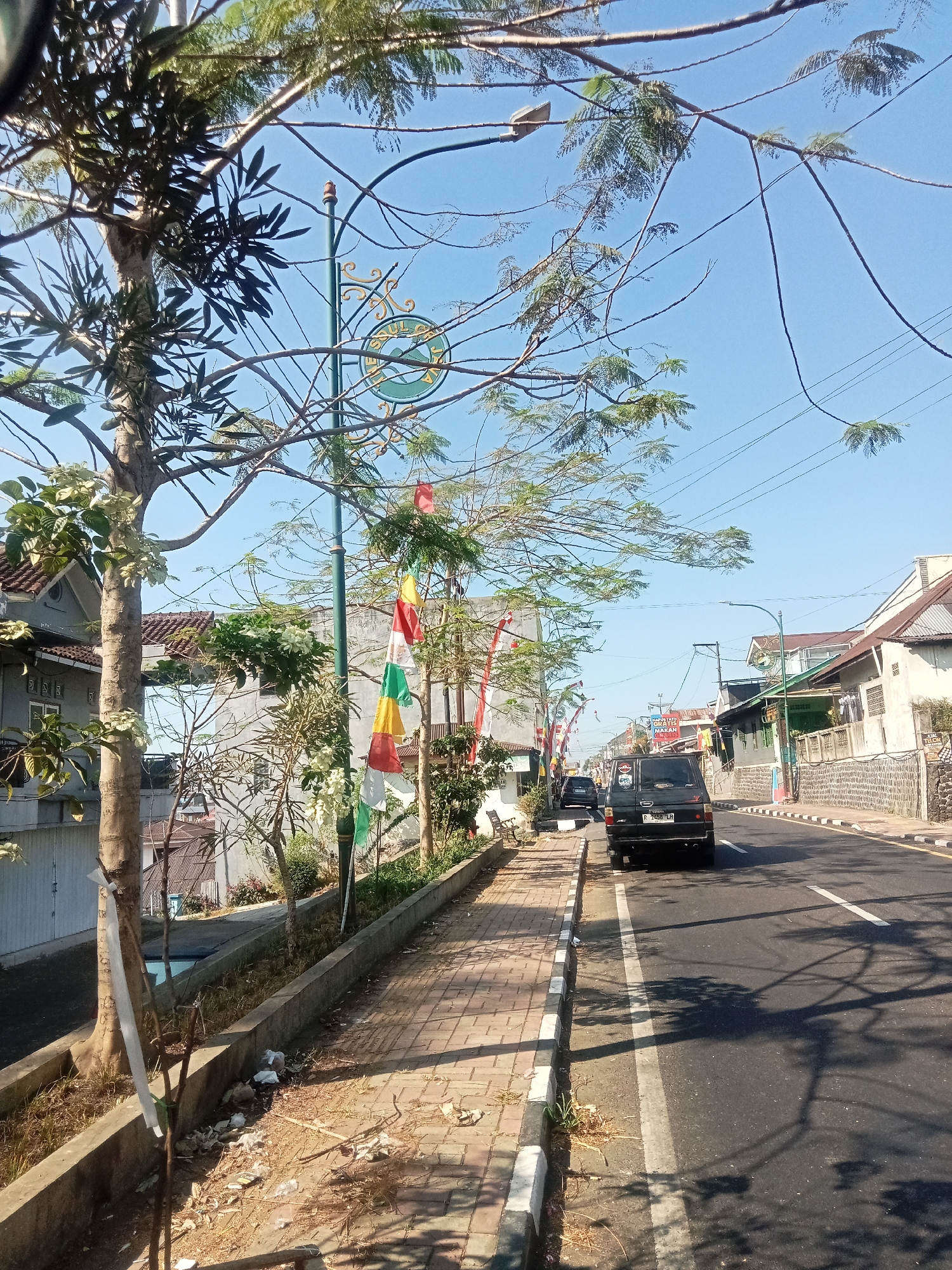 The image depicts a street scene in Jaraksari, Indonesia. A paved road runs through a moderately developed area with buildings on either side. A sidewalk runs along the left side of the road, bordered by a low concrete retaining wall and a strip of vegetation. Trees line the sidewalk, with some branches extending over the road.
Several vehicles are visible: a black SUV is parked on the right side of the road, and a black car is driving away in the distance on the left. The buildings appear to be a mix of residential and commercial, with some exhibiting traditional architectural elements. Flags of red and white, and other colors, are strung along the sidewalk. The sky is clear and blue, indicating sunny weather. Debris is scattered on the ground along the sidewalk and in the vegetated area. A street lamp is visible overhead. The overall impression is a typical street in a town or city in Indonesia.