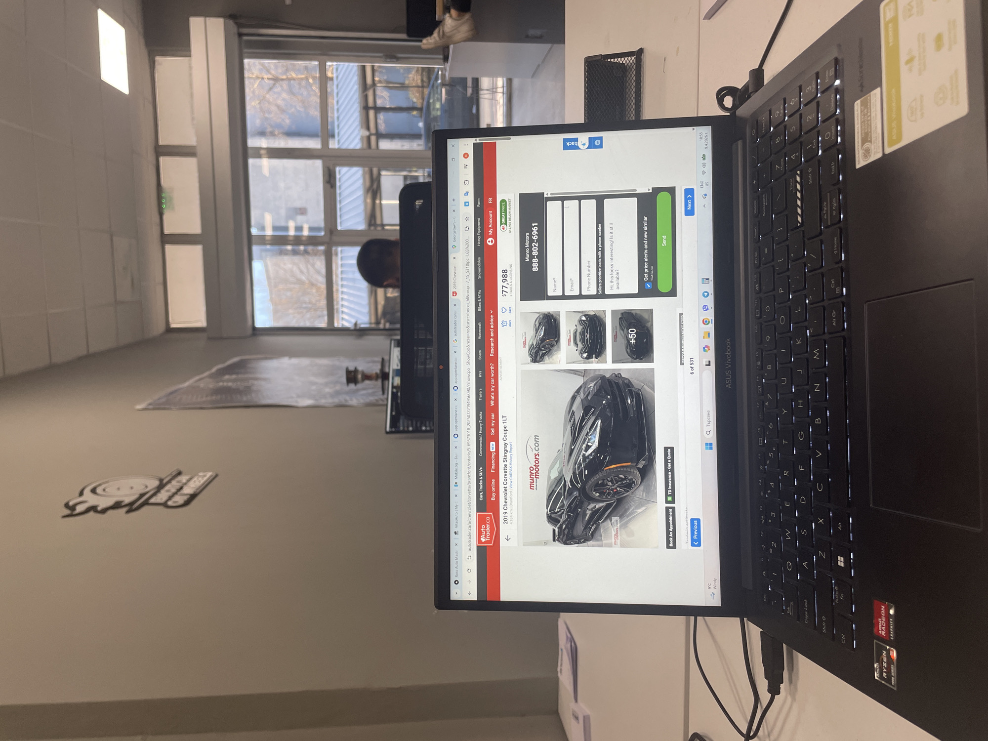 A close-up shot shows a black laptop computer displaying a web page from Autotrader.ca for a 2019 Chevrolet Corvette Stingray Coupe 1LT. The laptop's screen also shows a form to contact Munro Motors with a phone number of 888-802-6961, as well as tabs for Mobile.bg and Georgetown. A person is sitting behind the laptop, with a window visible behind the laptop. The logo "the spot" is on the wall. The location is not visible in the image.