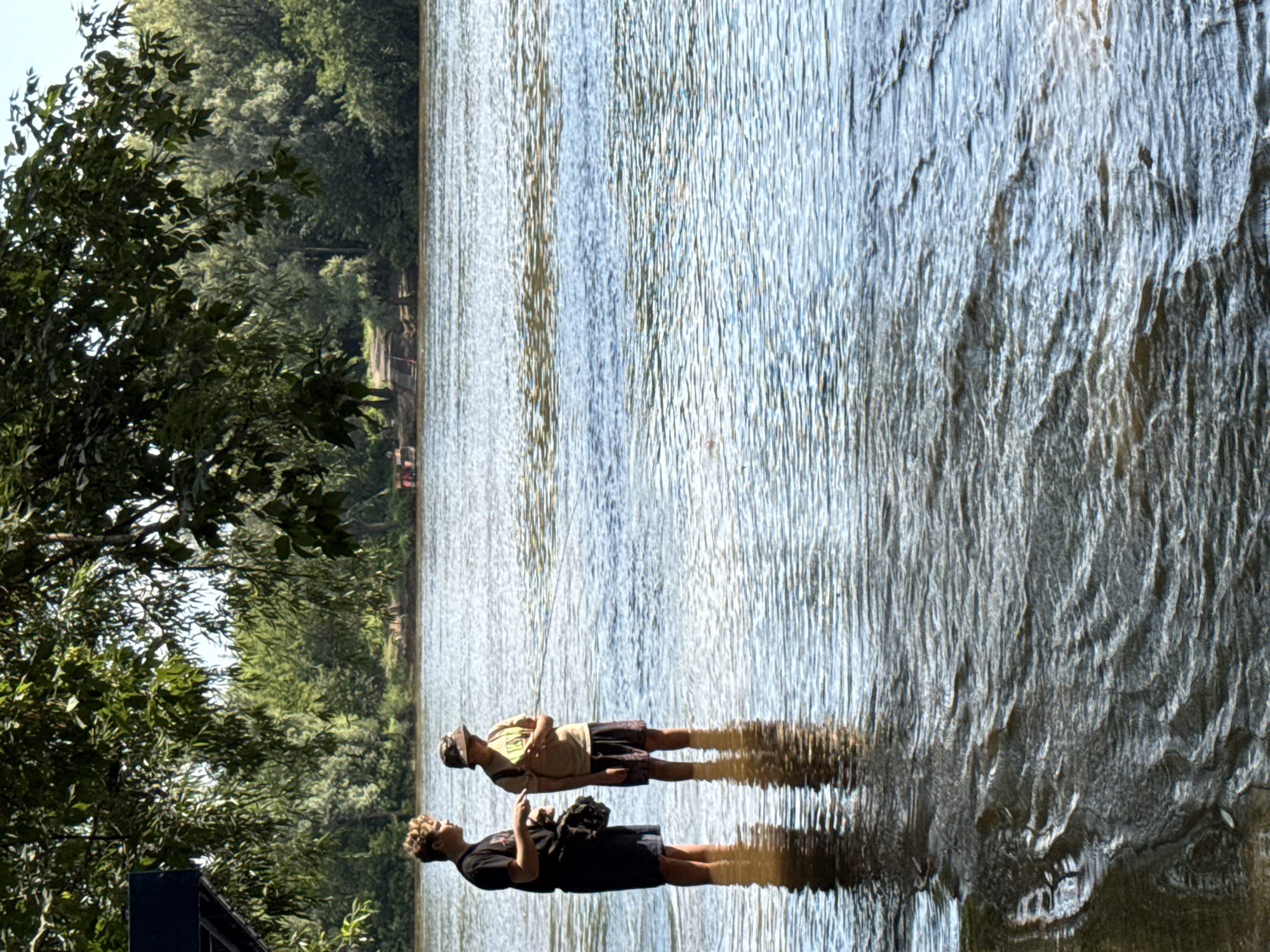 Two young males are standing in shallow, rippling water, possibly a river, under a tree with dense green foliage. The male on the left, wearing a black t-shirt and dark shorts, has light brown curly hair and is holding a black bag over his shoulder. The male on the right, wearing a light brown t-shirt, dark shorts, and a camouflage bucket hat, is holding a fishing rod, casting or retrieving. The background shows a treeline and a distant bank with some structures, suggesting an outdoor recreational area. The water has sunlight reflecting off its surface, creating a textured, shimmering effect. The scene is likely outdoors in a natural setting, possibly a park or riverside recreational area. Given the context of the prompt, this scene could be located along the Ufa River within the city of Ufa, Russia, a popular area for outdoor activities.