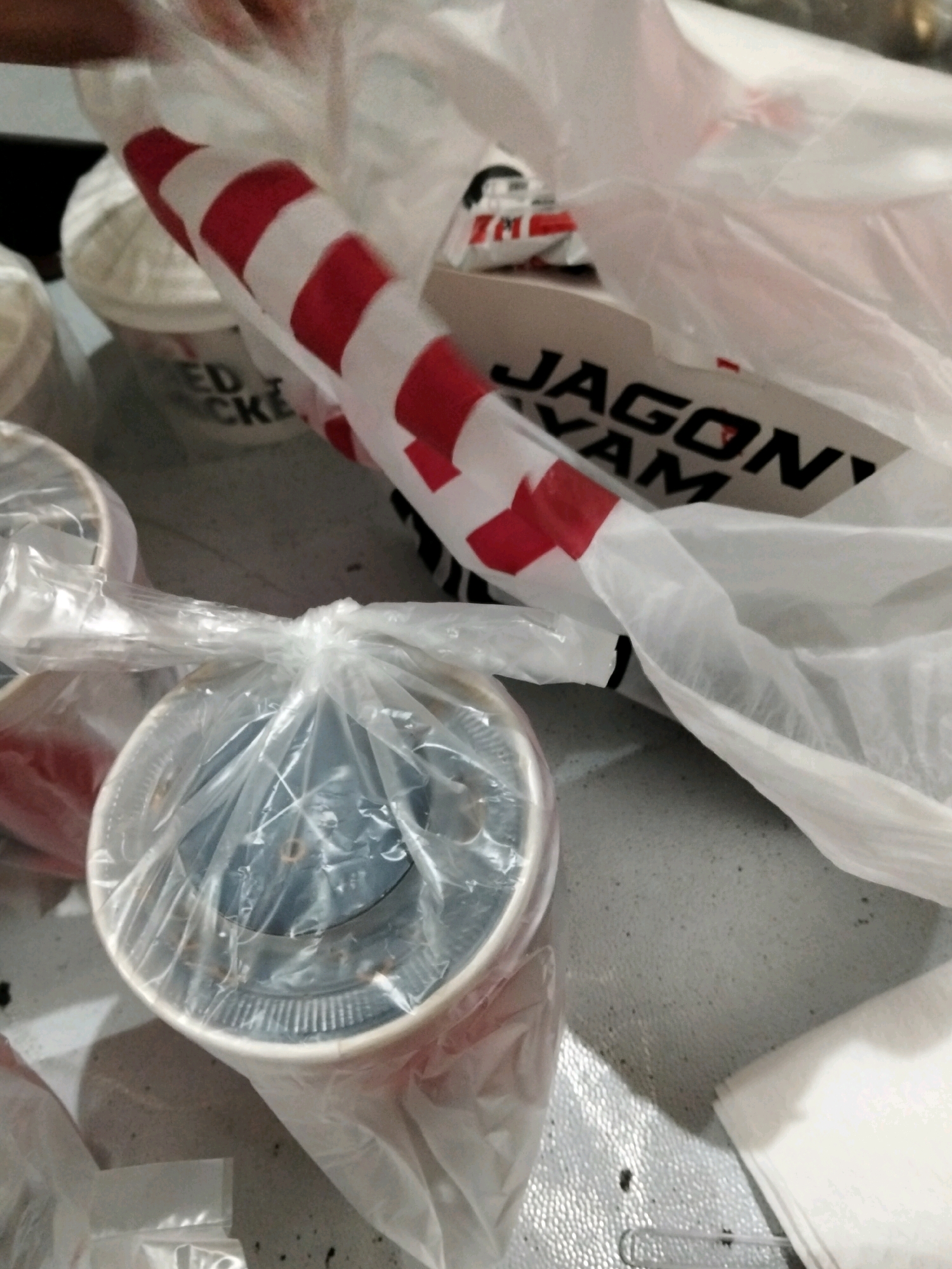 In Central Jakarta, Indonesia, this image captures a tabletop scene, likely inside a residence or establishment, showcasing several food containers and a plastic bag. Prominently featured is a white bucket with red and white stripes, partially obscured by a white plastic bag. Text on a white box within the bag reads "JAGON" and "YAM." Another white bucket, also wrapped in plastic, is visible in the foreground. A stack of white paper napkins rests on the right side of the table. The lighting suggests an indoor setting, possibly during nighttime or in a dimly lit area.