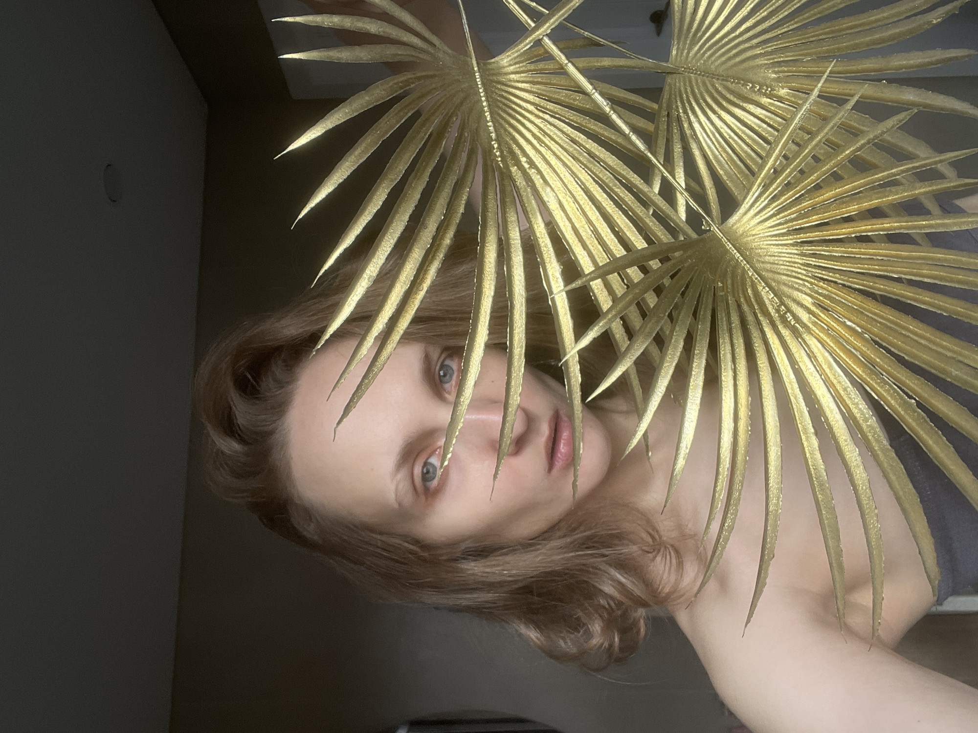 A woman with light skin and brown hair poses in front of a dark background. She is partially obscured by two gilded palm fronds. The location is unknown.