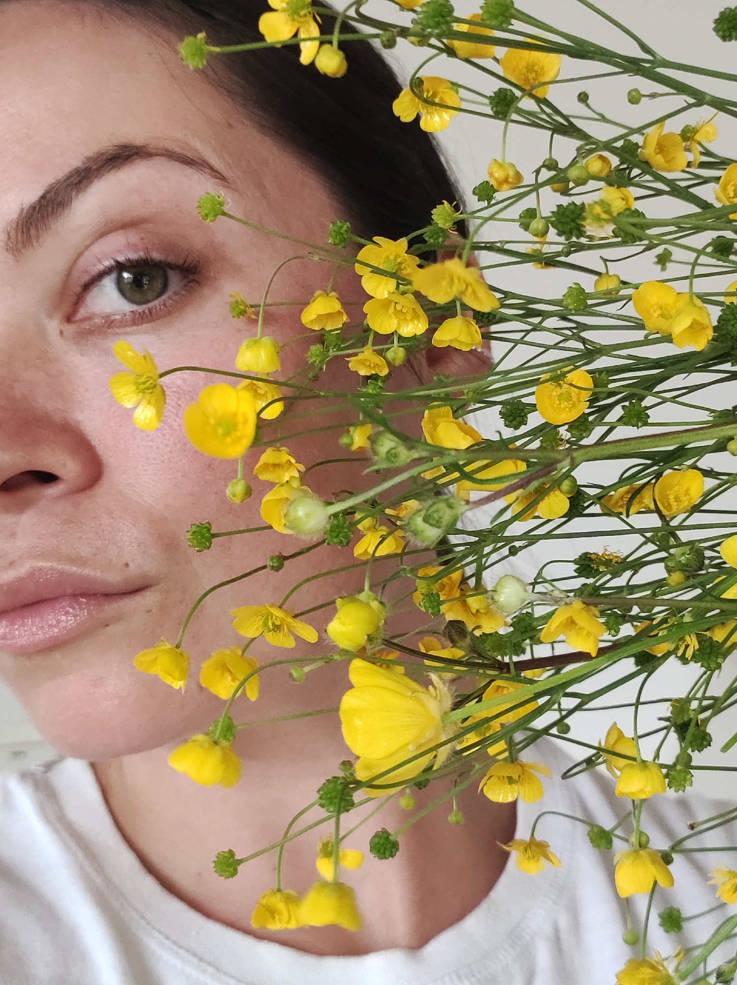A close-up, vertically oriented photograph features a woman partially obscured by a dense bouquet of bright yellow flowers. The left side of the frame shows a portion of the woman's face, including her left eye, eyebrow, a section of her nose, cheek, and lips. Her dark hair is visible on the upper left, and she appears to be wearing a white t-shirt or top. Her left eye, a shade of green or hazel, is open, with a visible pupil and eyelashes, conveying a calm or neutral expression. Her lips are slightly parted.
The right side of the image is dominated by numerous small, vibrant yellow flowers, likely buttercups, with slender green stems and some green buds. These flowers extend across her face, creating an artistic and natural composition. The background is a plain, uniform light color, suggesting an indoor setting with diffused lighting. The soft illumination highlights the natural texture of her skin and the delicate details of the flowers. There is no visible text. The overall mood is serene and highlights natural beauty.