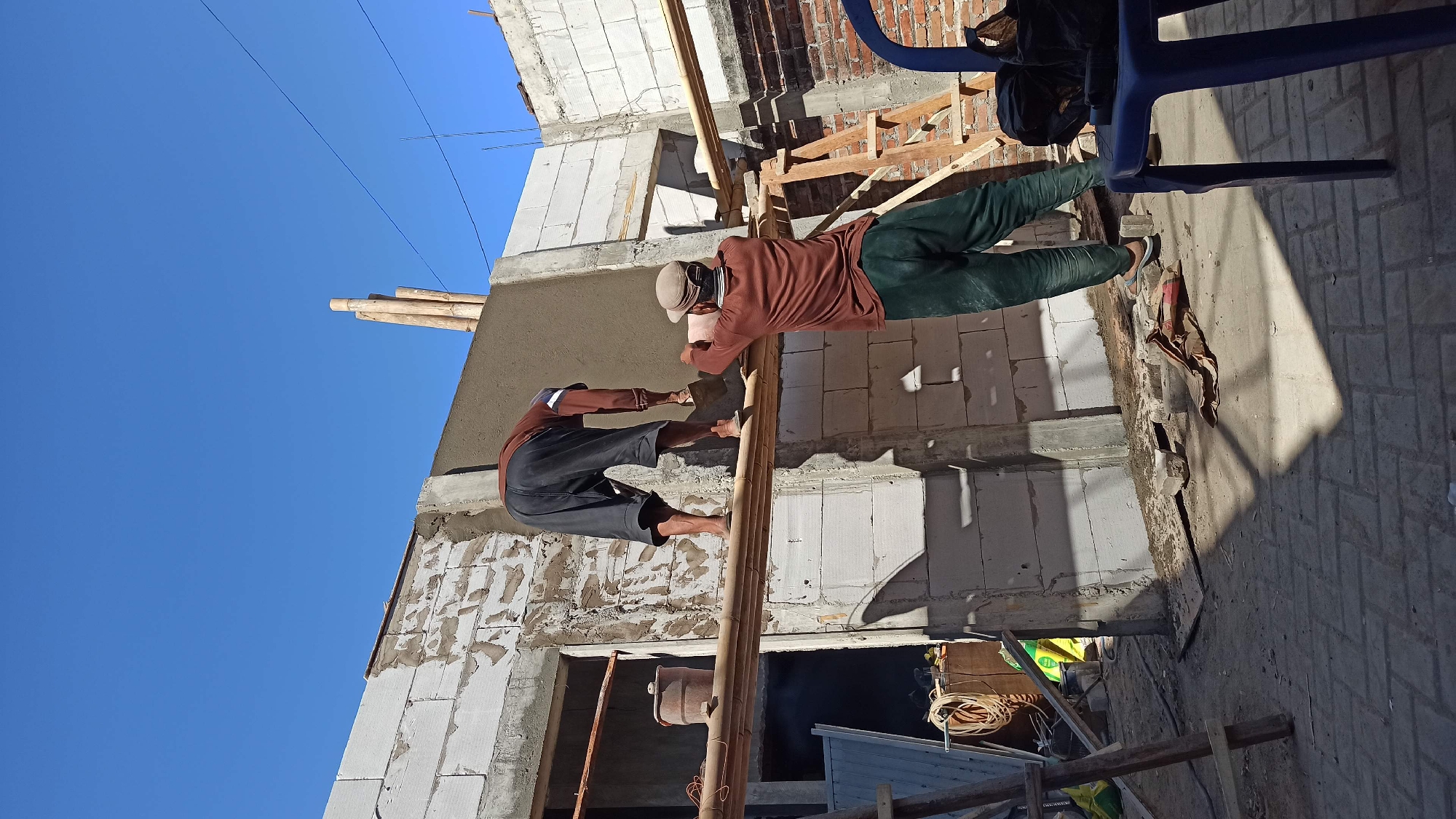 Two men are plastering a wall outdoors, likely in the city of Ngumpul, Indonesia, under a clear blue sky. The man on the left, wearing a brown long-sleeved shirt and dark shorts, is bent over and applying plaster to a white block wall with a trowel. He is standing on a horizontal bamboo pole supported by wooden scaffolding. The man on the right, facing away from the camera and wearing a brown long-sleeved shirt, dark cap, and green pants, is also applying plaster to the same wall, using a trowel and standing on a similar bamboo pole. The construction site features white cinder blocks, cement, and various construction materials. Shadows cast by the bamboo scaffolding and structures are visible on the ground, which is paved with interlocking bricks. A blue plastic chair and a black bag are to the right of the frame.