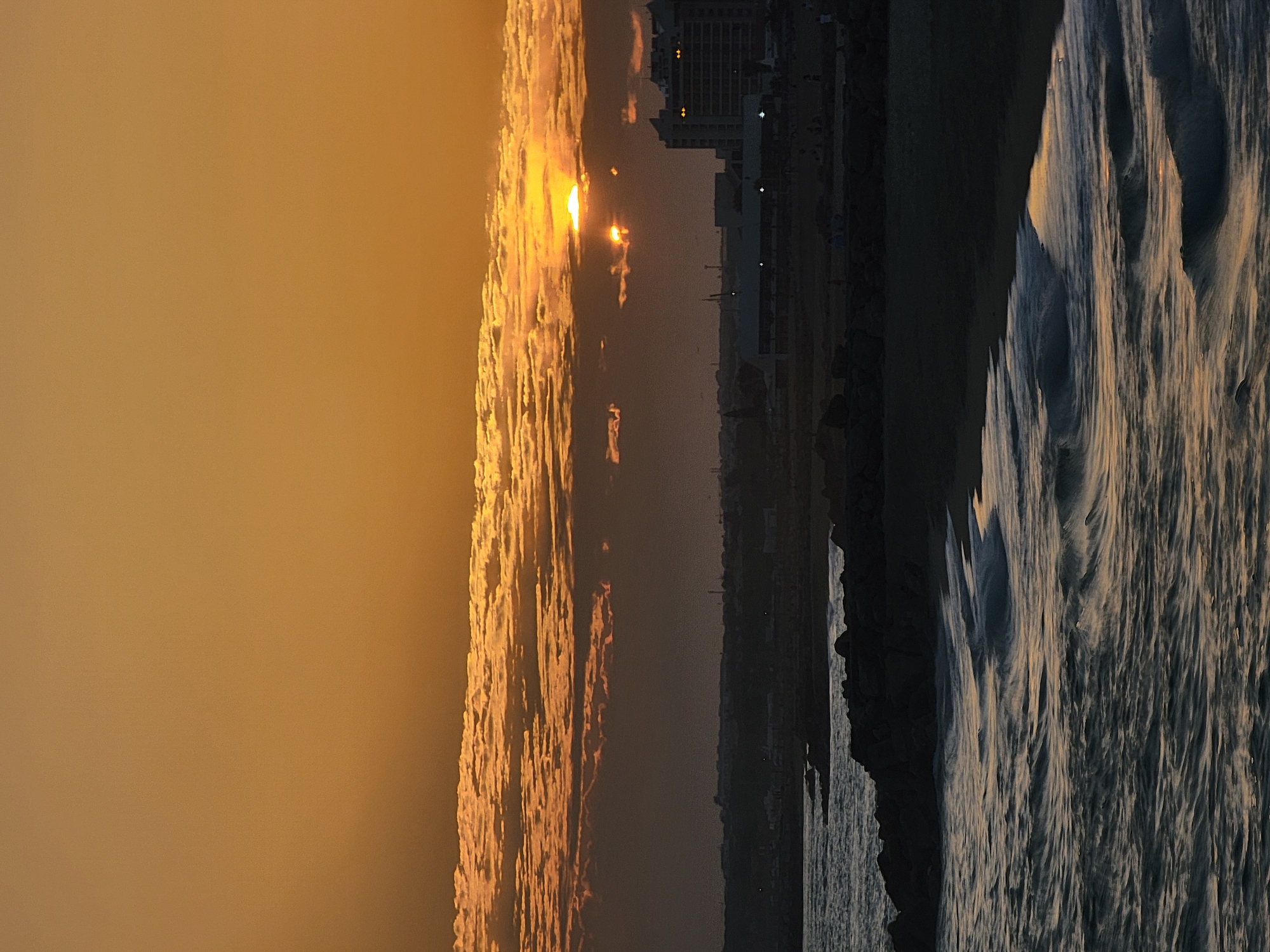 The image depicts a serene sunset over the ocean in Quarteira, Portugal. The sky is ablaze with warm orange hues, with streaks of sunlight piercing through scattered clouds. The sun, partially obscured by clouds, casts a golden glow on the water, which laps gently against a dark, rocky shore and a sandy beach. In the background, the silhouette of the city of Quarteira is visible, with buildings and distant lights suggesting a vibrant coastal town. A few figures can be seen on the beach, enjoying the tranquil evening. The overall atmosphere is one of peace and natural beauty.
