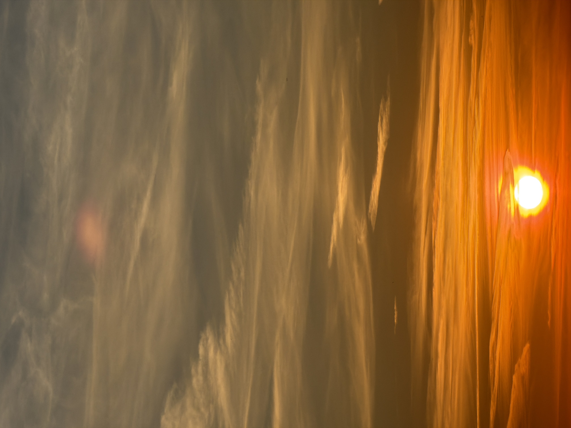 The media file captures a panoramic, vertically oriented view of a sky during what appears to be either sunrise or sunset, characterized by a vibrant and dramatic color palette. The dominant subject is the sky filled with streaky, elongated clouds and a prominent sun.
On the far right side of the image, a very bright, glowing sun is visible as an intense white-yellow orb, partially obscured by atmospheric conditions or thin clouds. Its powerful light creates a strong lens flare and bathes the adjacent sky and clouds in brilliant orange and red hues.
The sky is largely covered by wispy, cirrus-like clouds that stretch across the frame. These clouds catch the low sunlight, appearing golden yellow, orange, and reddish near the sun, and gradually fading into more muted grays, browns, and faint blues towards the left side of the image where the light is less direct. The backlighting highlights the intricate textures and forms of the clouds, giving them an ethereal quality.
The scene conveys a sense of natural beauty and vastness, typical of a twilight period. There are no visible landscape features, buildings, people, or identifiable objects other than the sky and clouds. A tiny, indistinct dark speck is noticeable in the mid-left area of the sky in one of the magnified views, but it is too small to identify definitively. No text is visible in the image.