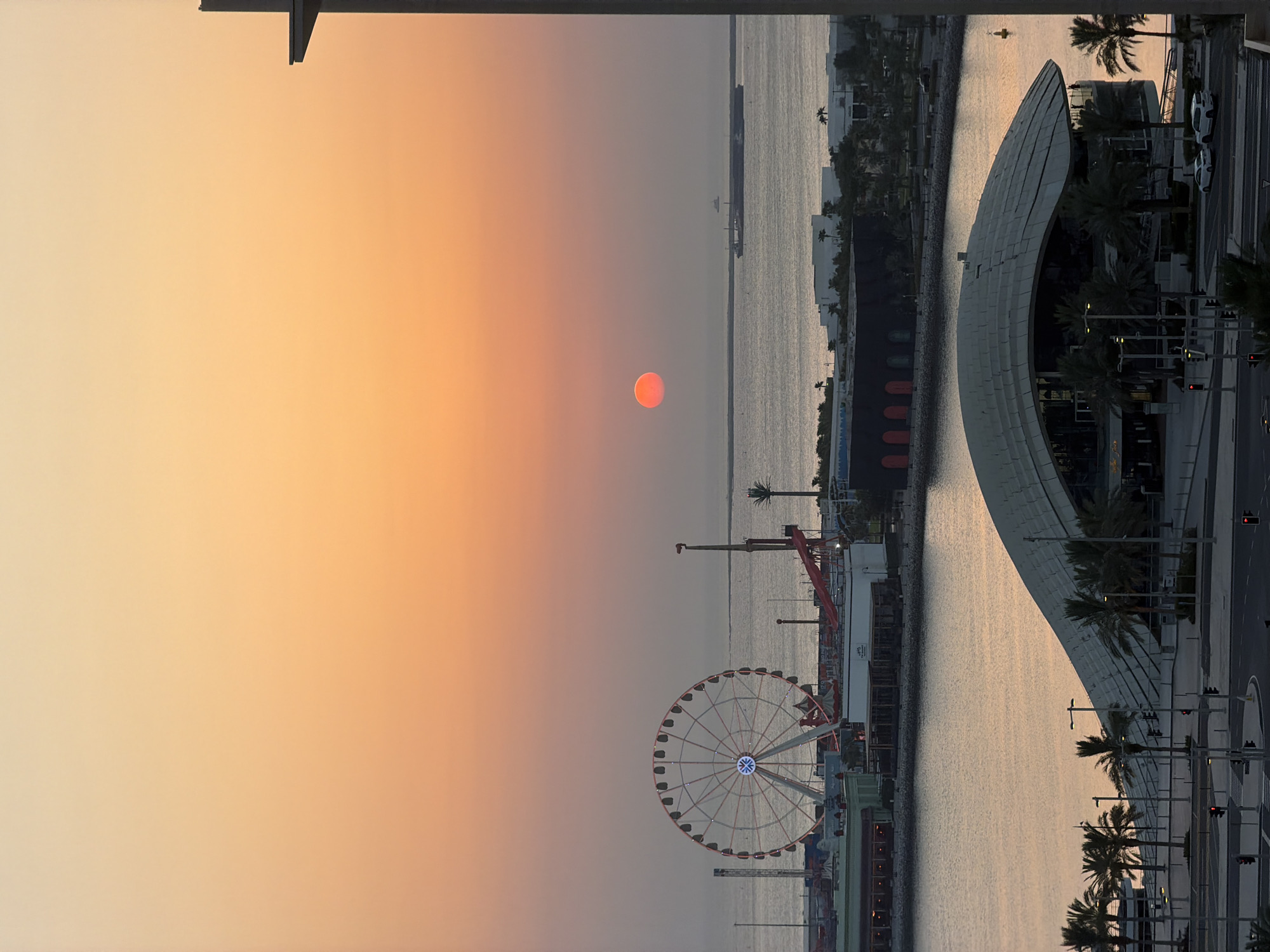 This is a vertical, medium-shot, high-angle photograph of a coastal city at sunset. The sky is a gradient of soft orange, transitioning to a lighter peach hue towards the top. A large, circular Ferris wheel with illuminated spokes dominates the left side of the middle ground. Adjacent to it, other amusement park rides are visible, including a tall, slender tower ride with red accents. The middle ground also features a body of water, a pier, and several low-rise buildings. In the distance, a large cargo ship and a smaller vessel are visible on the horizon.
The foreground of the image features a modern, architecturally distinctive building with a curved, metallic roof, reminiscent of an amphitheater or convention center. Palm trees are scattered around this building and the surrounding paved area, which includes roads and streetlights. The water in the foreground reflects the warm tones of the sunset. A sliver of a dark vertical structure is present on the right edge of the frame, suggesting the viewpoint is from a building overlooking the scene. Two white cars are parked on the road in the foreground. The overall impression is that of a lively waterfront area, likely a tourist destination, bathed in the warm glow of a setting sun.