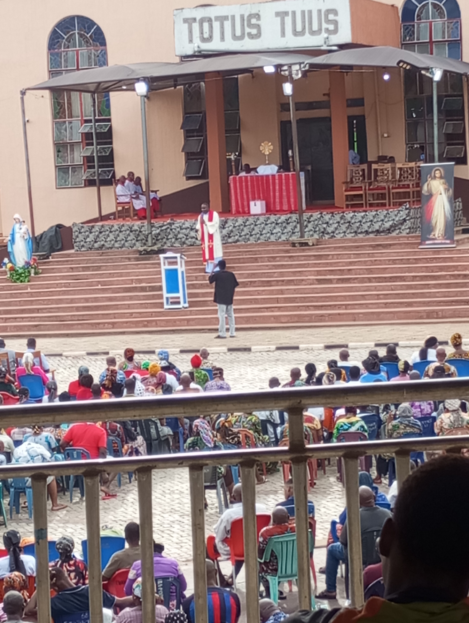 The image shows a religious gathering in Onitsha, Nigeria, at a location with the words "TOTUS TUUS" displayed prominently on a building. A large crowd of people is seated outdoors in plastic chairs, facing a stage area with several levels of steps. On the stage, there are individuals in religious attire, including a priest in a white and red robe who is speaking from behind a blue podium. A table covered with a white and red cloth sits behind the priest. There are other people seated on the stage, and a standing banner with an image of Jesus is present. A statue of the Virgin Mary is placed on the left side of the steps leading up to the stage. The image is taken from behind a barrier, possibly a metal railing, with a person's head partially visible in the lower right corner of the frame. The weather appears to be daylight.