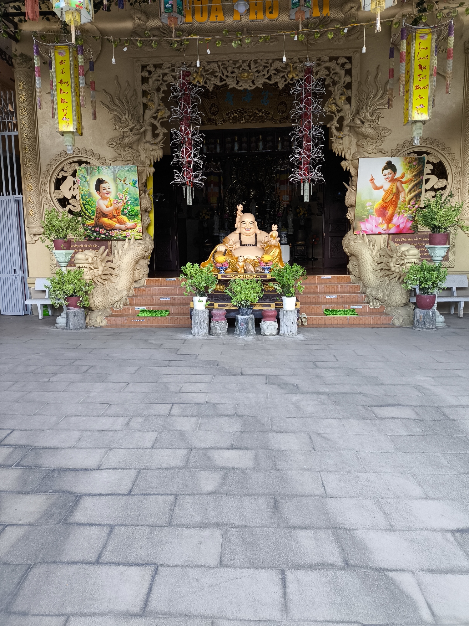 This is a photograph of a Buddhist temple entrance in Bac Nha Trang Ward, Vietnam. The scene depicts a tranquil setting with several religious elements. In the foreground, a paved courtyard with large, grey stone tiles leads up to a set of stairs. The stairs are adorned with decorative dragon sculptures on either side, which are a common motif in Vietnamese temples.
At the top of the stairs, a large, golden statue of a smiling Buddha is prominently displayed, seated with his hands resting in his lap. To the left of the Buddha statue, a painting shows a young boy in orange robes holding a flower, with birds nearby. To the right of the Buddha, another painting shows a similar figure, also in orange robes, standing on a lotus flower, with his hand raised in a gesture of blessing. Both paintings appear to be depicting Budai, also known as the Laughing Buddha, or a young Siddhartha.
The entrance to the temple itself is recessed and appears to be dark, suggesting an interior space. Hanging from the ceiling above the entrance are ornate lanterns and banners with Vietnamese text. The overall atmosphere is one of peace and reverence. The lighting suggests it is daytime, and the weather appears to be fair, as there are no signs of rain or strong sunlight. The scene is characteristic of a place of worship, inviting contemplation and spiritual reflection. The visible text on the banners likely contains religious verses or blessings.