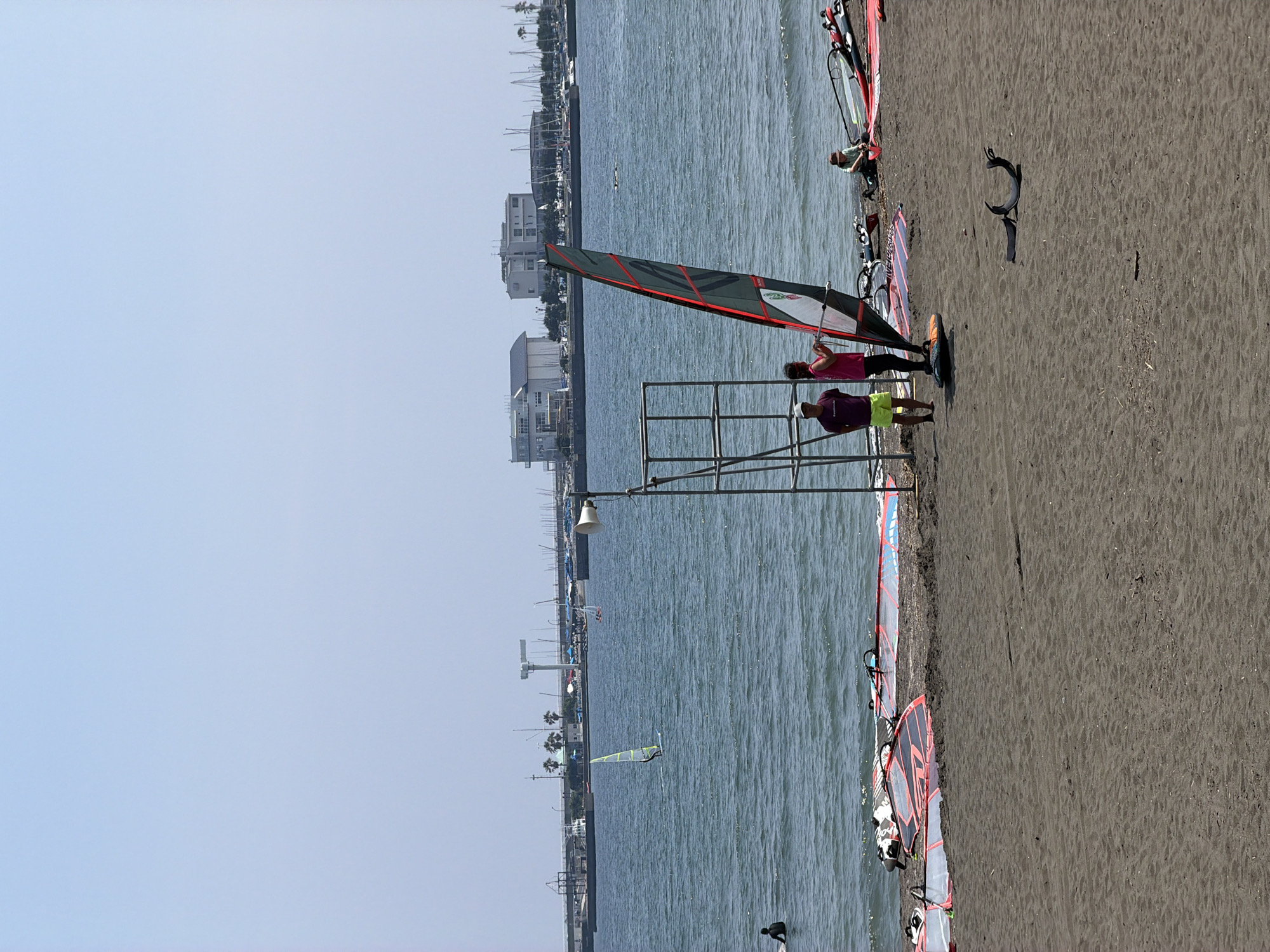 This image captures a scene at the beach in Fujisawa, Japan, likely near Katase Kaigan or Enoshima, known for water sports. The sky is clear and pale blue, indicating a sunny day. The water is a calm, dark blue with gentle ripples.
In the foreground, a sandy beach is visible, with several windsurfing boards and sails laid out on the sand. The boards are colorful, featuring designs of red, blue, and white.
On the beach, near a metal structure that appears to be a lifeguard stand or observation post, two people are standing. One person, wearing a purple shirt and yellow shorts, is looking towards the water. Beside them, a person in a pink top and dark pants is holding onto a windsurfing sail, which is attached to a board. Another person is seated further down the beach, near some windsurfing equipment. In the water, a lone windsurfer with a yellow sail is actively riding the waves. Further out, another windsurfer can be seen.
In the background, the coastline features buildings, some appearing to be white and modern, and others with darker roofs. There are also palm trees and a harbor with numerous sailboats and yachts docked. A large crane structure is also visible on the waterfront. The overall atmosphere is serene, with a focus on recreational water activities.