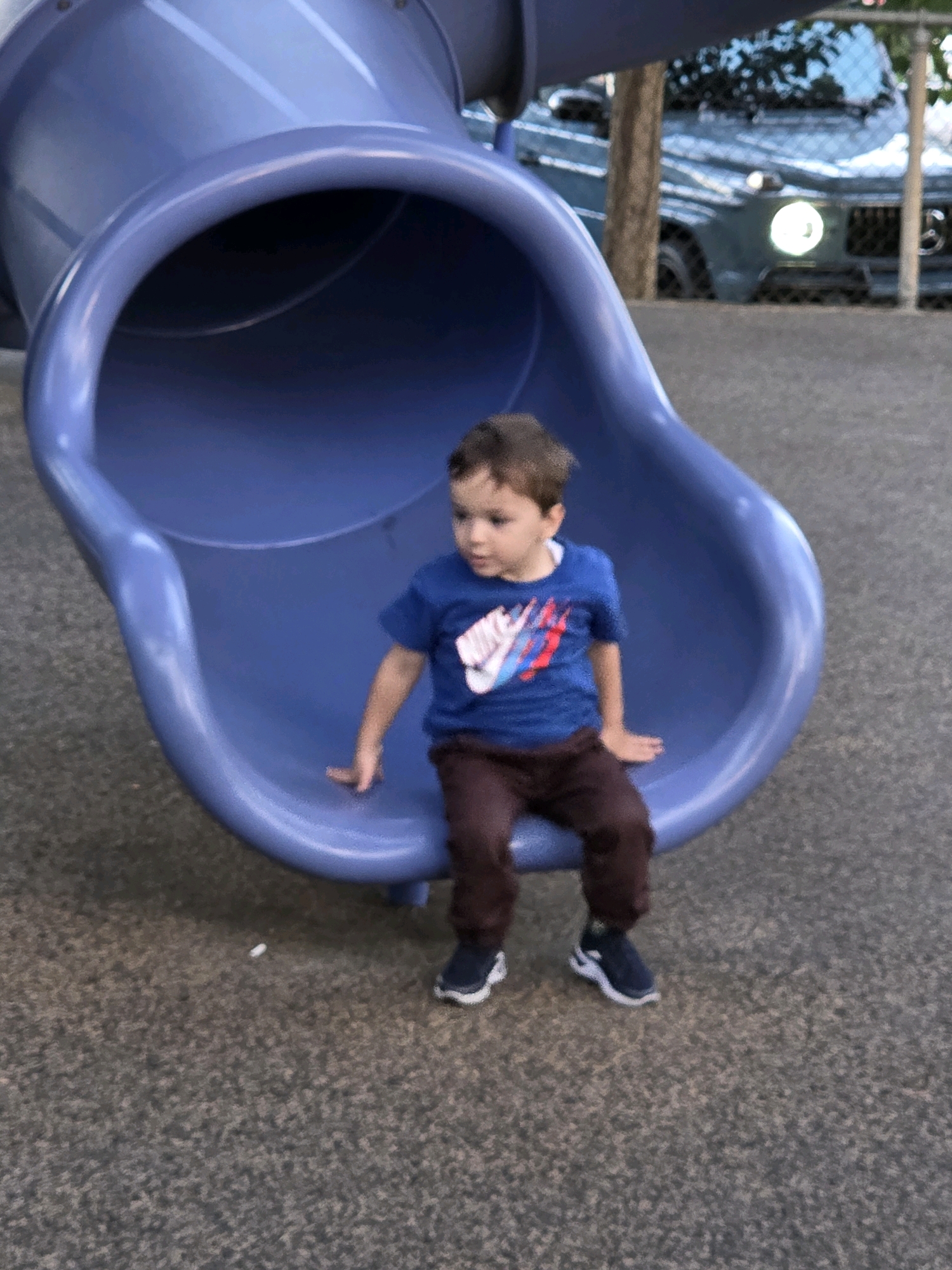 A young boy is sitting on the edge of a large, blue plastic slide in a park. He is wearing a blue t-shirt with a red and white logo on it, dark pants, and dark sneakers with white soles. The slide is curved and open at the bottom, with a textured gray surface on the ground around it. In the background, a dark-colored SUV is parked, partially obscured by a tree and a chain-link fence. The lighting suggests it is daytime.