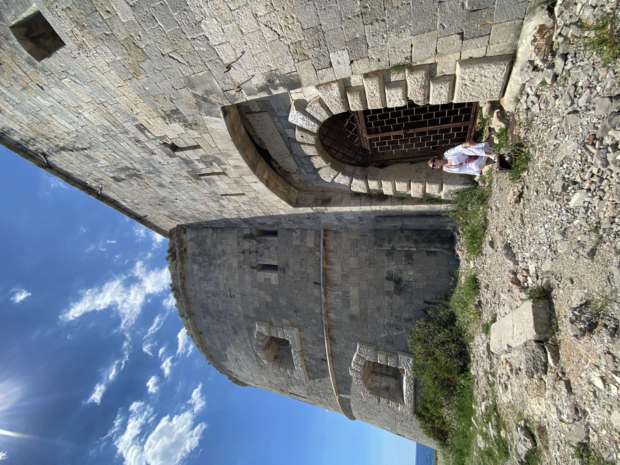 A low-angle, slightly tilted outdoor shot depicts a stone fortress under a partly cloudy blue sky. The fortress wall is constructed from light-colored stone blocks. The lower section of the fortress has several rectangular embrasures with decorative stonework. The upper part of the fortress appears to overhang, with a barred, arched opening near the top, constructed from stone blocks.
A person wearing white clothing sits on the rocky and grassy ground next to the fortress wall, appearing to be looking into the arched opening. Vegetation clings to the side of the fortress and the surrounding ground.
The location is likely on the Luštica peninsula in Montenegro, given the type of stone structure, landscape, and geography.