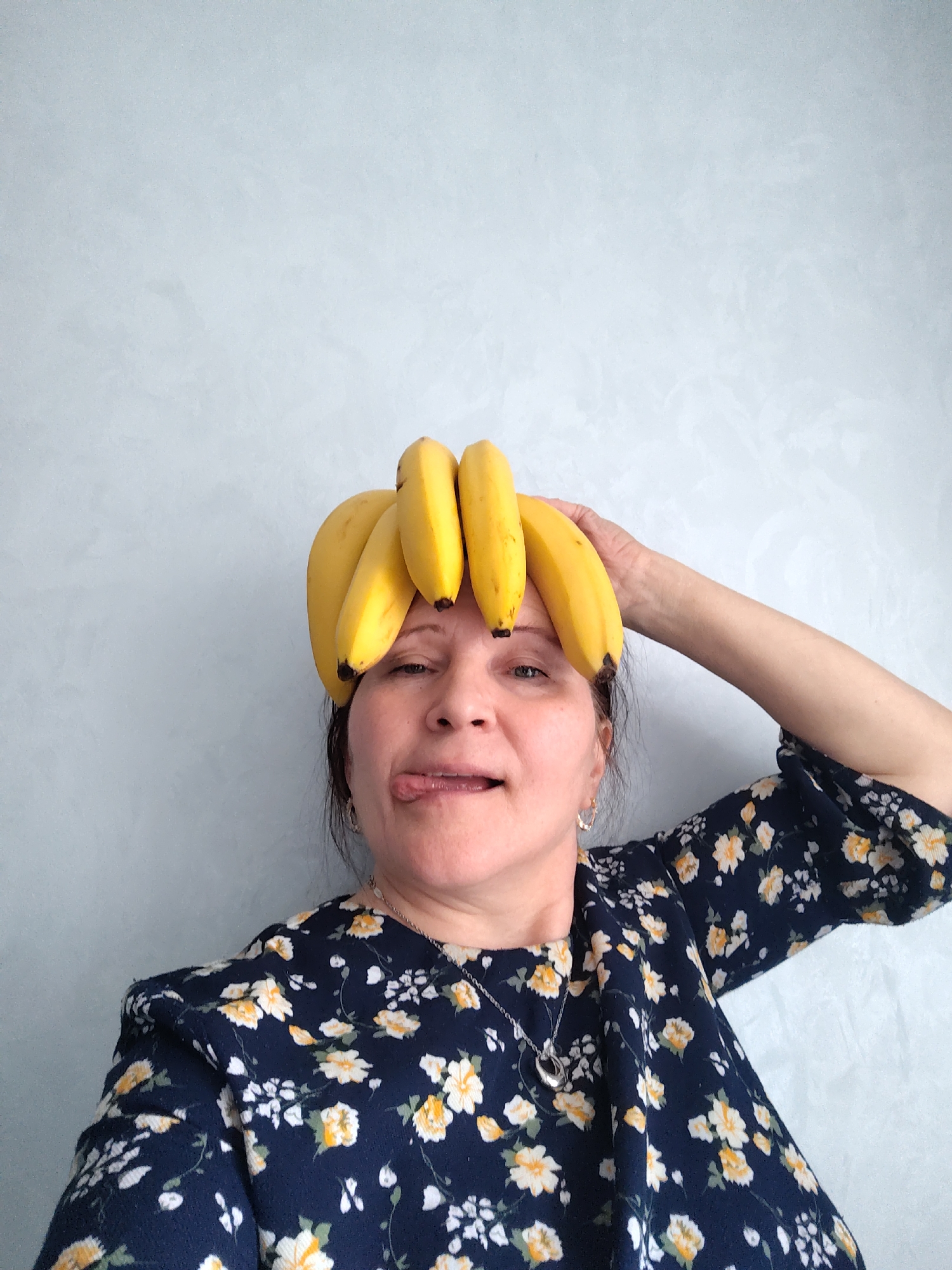 A woman is posing for a selfie indoors against a light grey textured wall. She has a bunch of yellow bananas placed on her head. Her right hand is raised, holding the bananas in place. She is wearing a dark blue, floral print dress and a silver necklace. She is sticking her tongue out, making a playful expression. The location is not specified.