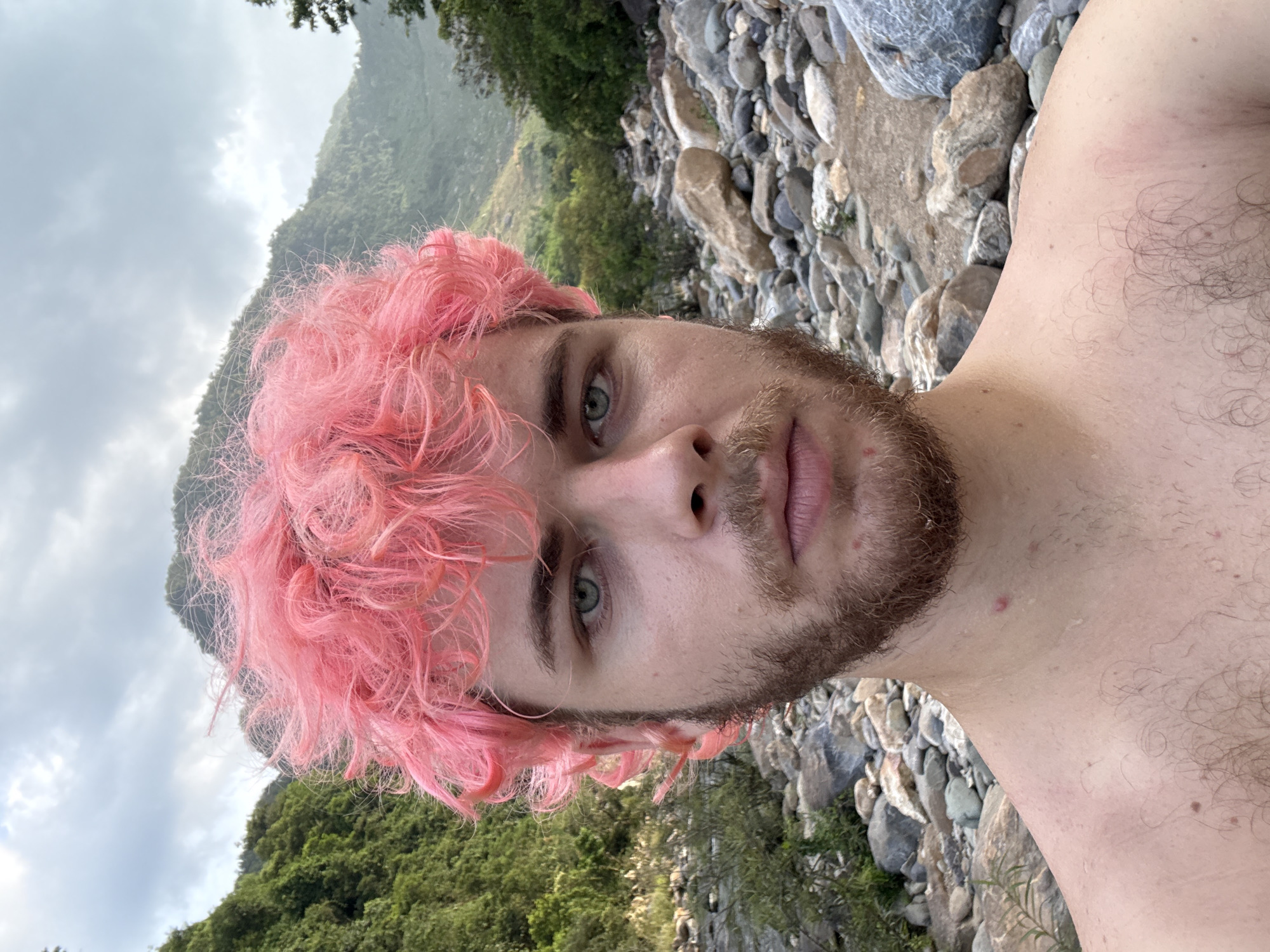 A close-up, angled selfie depicts a man with pink curly hair, a beard, and light eyes, shirtless, seemingly outdoors. The background features a rocky area with vegetation and a mountain. There is no specific information to pinpoint the location in Đà Nẵng, Vietnam, from the image alone.