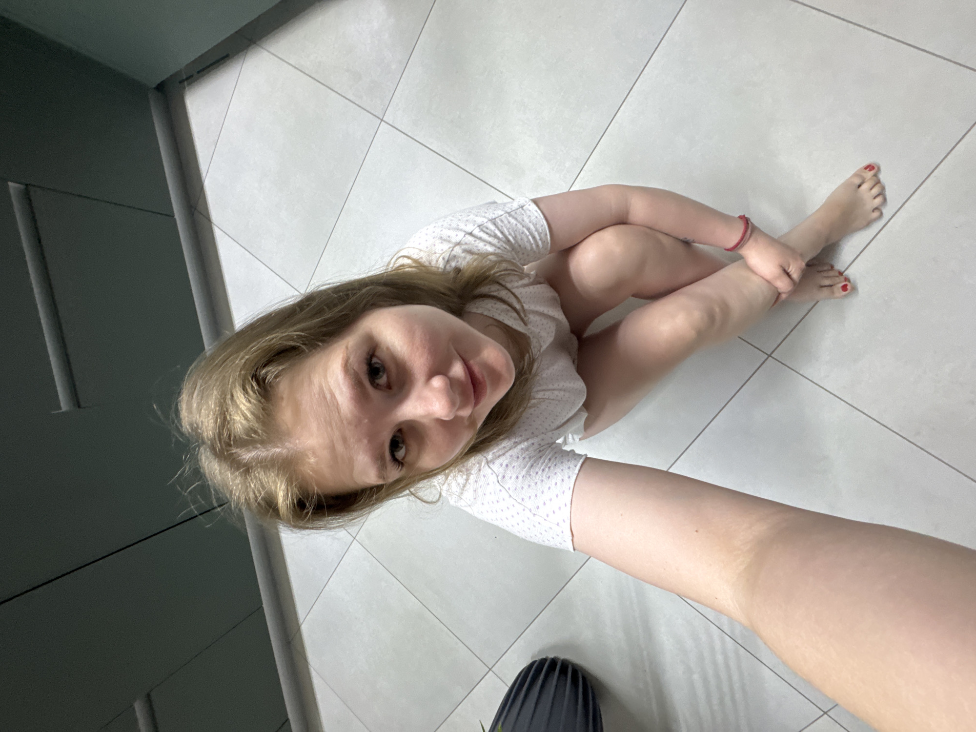 A young woman with blonde hair is sitting cross-legged on a tiled floor, looking up at the camera. She is wearing a white, short-sleeved shirt with small patterns. Her toes are painted red and she has a red bracelet on her ankle. Part of a black object with ridges is visible in the lower left of the frame. The floor appears to be a bathroom or kitchen. The location is in a room with light-colored tiles somewhere in the city of Gornyi (Dvurechenskoe S/P), Russia. The image is taken from a high angle, possibly as a selfie.