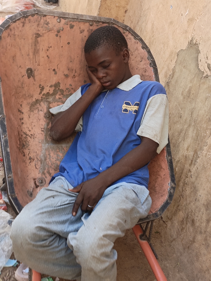 A young individual, identified as male, is depicted seated within the orange-red, heavily rusted metal tub of a wheelbarrow, which functions as a makeshift chair. The scene is located in Kawaji, Nigeria. The male, with dark skin and short black hair, is positioned with his eyes closed, head resting on his right hand, suggesting sleep or deep rest. He wears a short-sleeved blue polo shirt featuring light beige/off-white sleeves and collar, with a yellow "M" logo and "AMARSHA" text on the left chest. Light grey trousers are worn, and a silver-colored ring is visible on the ring finger of his left hand. The immediate background to the right consists of a light brown wall exhibiting wear and possible damage. The ground beneath the wheelbarrow shows scattered debris, likely plastic materials, on a dirt surface.