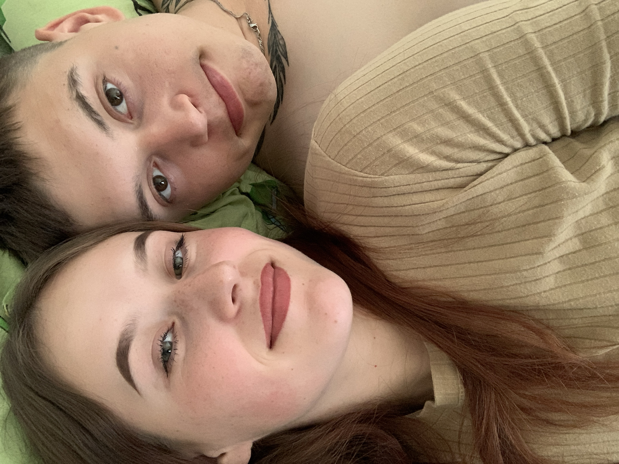 Two people lie close together, face-up. The person on the left has short dark hair, brown eyes, light skin, and a light-colored shirt. A tattoo is visible on the upper left chest. The person on the bottom has light skin, brown hair, green eyes, freckles, and is wearing a brown-colored top. A green surface is beneath them. The environment is indoor. There is no evidence to support the location being Фоки, Russia.