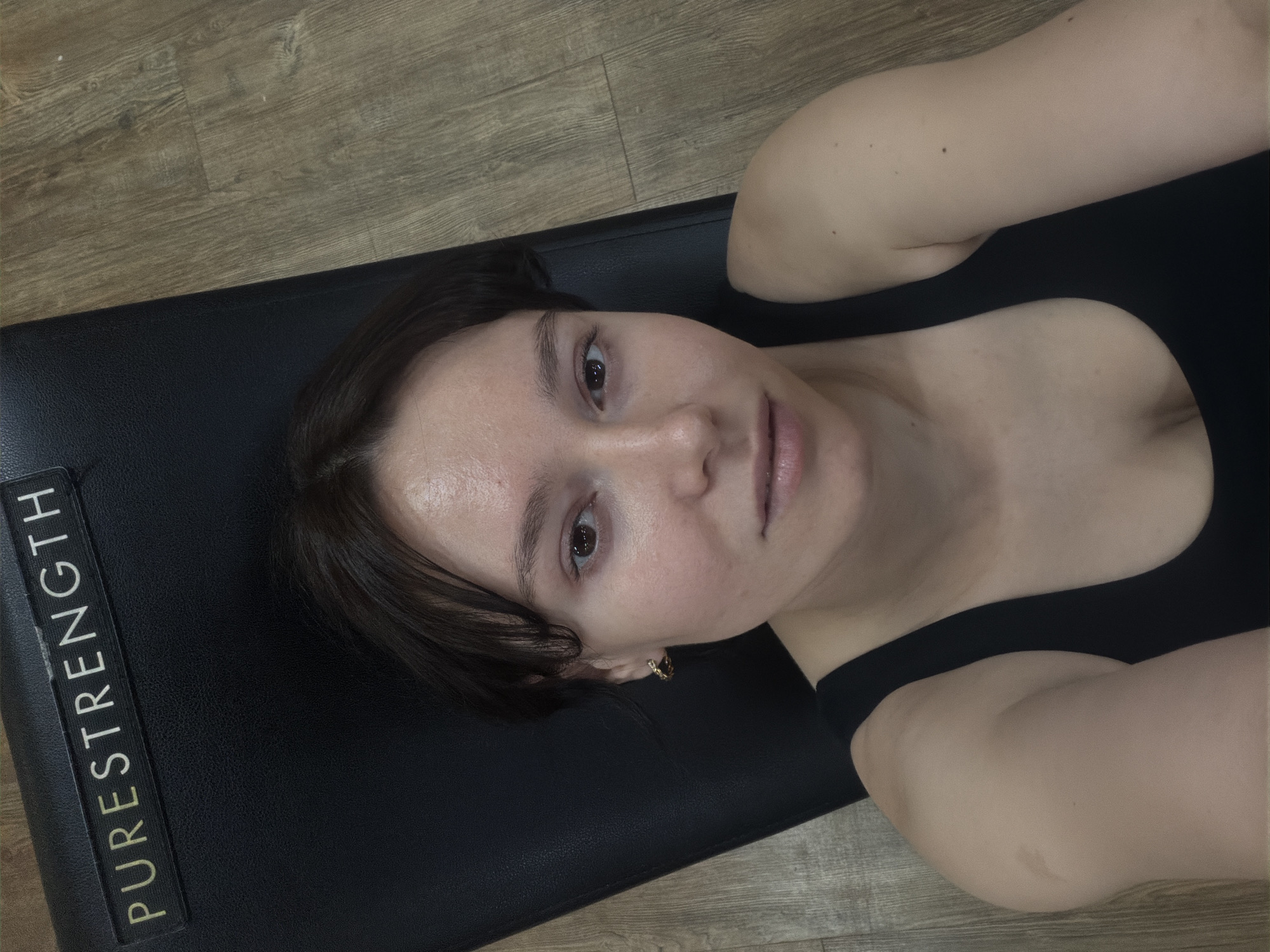 A young woman with light skin and dark brown hair is lying on a black gym bench. She is wearing a black tank top and a small gold hoop earring in her left ear. Her head is tilted slightly to her left, and she is looking directly at the camera. The gym bench has a gold and black "PURESTRENGTH" logo on it. The background consists of light brown wooden floorboards. The scene appears to be indoors, in a gym setting. The location is Chaikovsky, Russia.