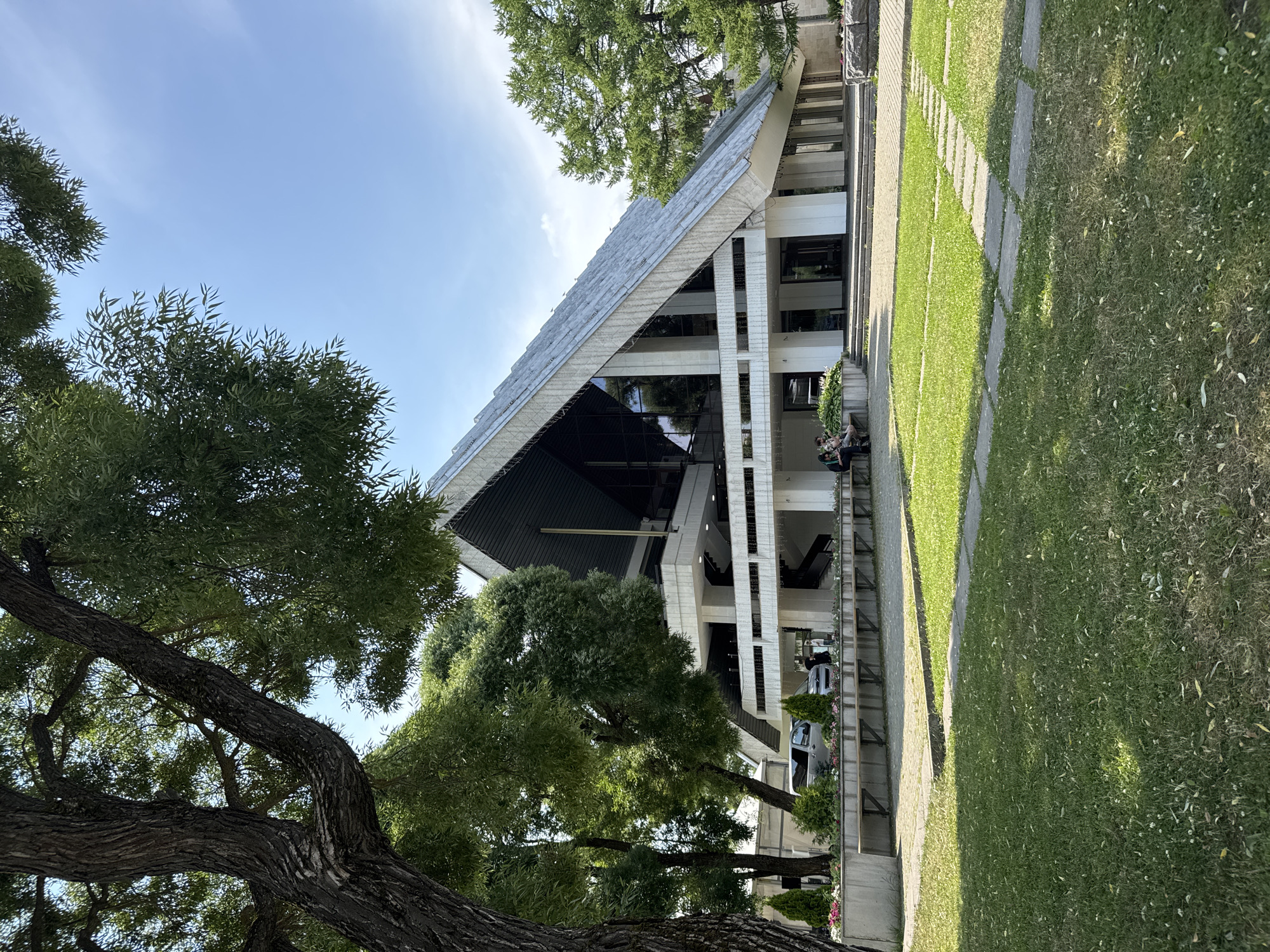 A modern, angular building with a large, sloping roof is the central focus of the image. The building appears to be made of concrete and glass, with multiple levels and large windows. It is surrounded by greenery, including large trees and a well-maintained lawn. Several people are visible, some sitting on benches and others standing near the entrance. The sky is clear and blue, suggesting a sunny day. The scene is quiet and serene, with no obvious signs of activity.