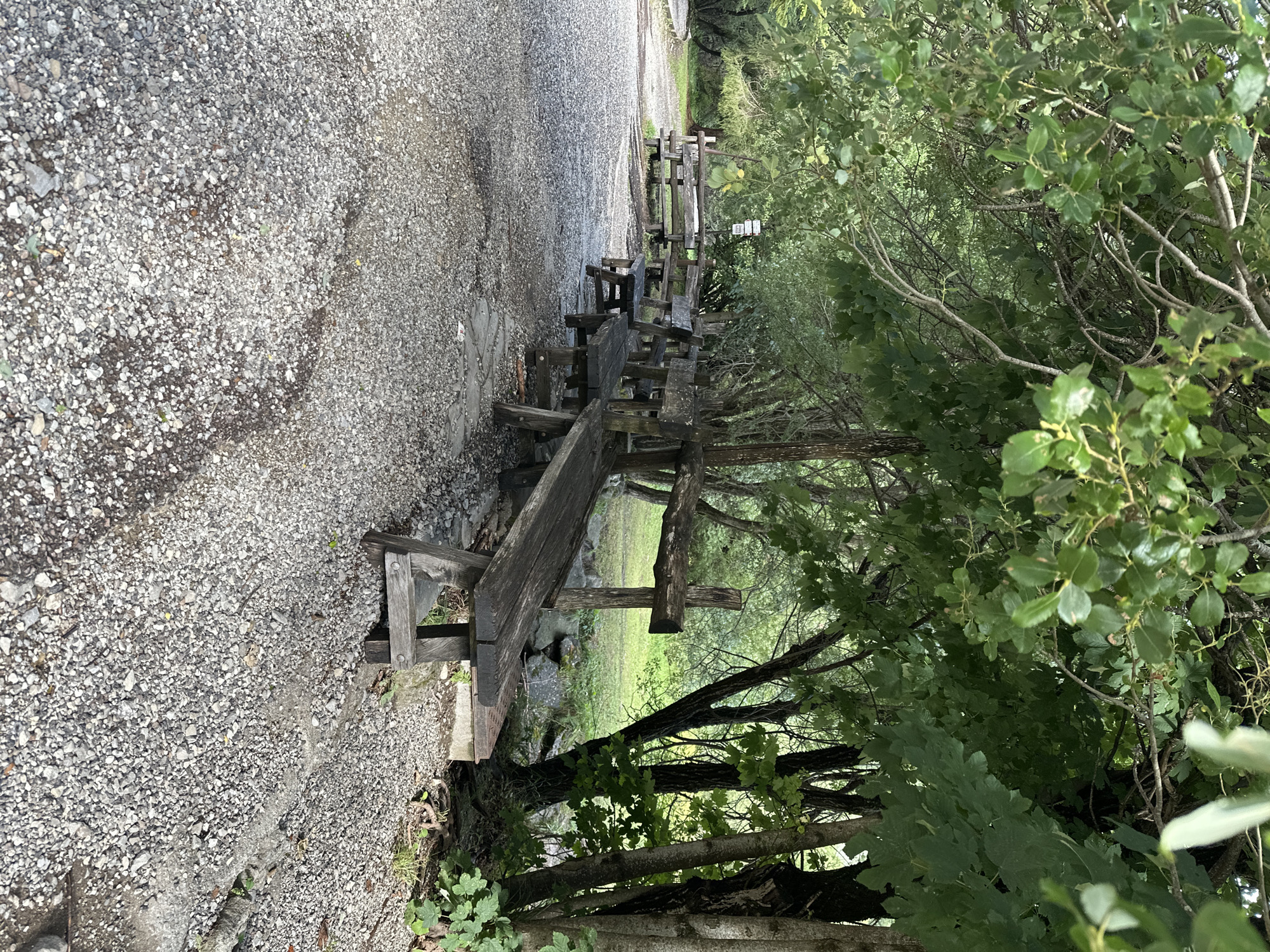 The image shows a rural outdoor scene in Corniglio, Italy, featuring a gravel path lined with weathered wooden benches under a canopy of green trees. The path is covered in small, light gray stones. Several rustic wooden benches with thick plank seating and legs are arranged along the path, some appearing to be a picnic table setup with benches. In the background, more trees and foliage create a natural, shaded environment. A wooden fence is visible behind one of the benches. The overall impression is of a quiet, possibly recreational area within a wooded setting. There are no people visible in the image.