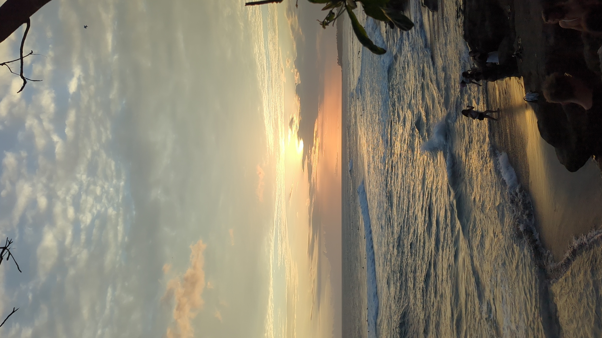 The image shows a sunset view of the beach and ocean in North Kuta, Indonesia. The sun is setting over the ocean, casting a golden glow on the water and sand. People are visible along the beach. The sky is filled with clouds. Tree branches are visible in the top left corner.