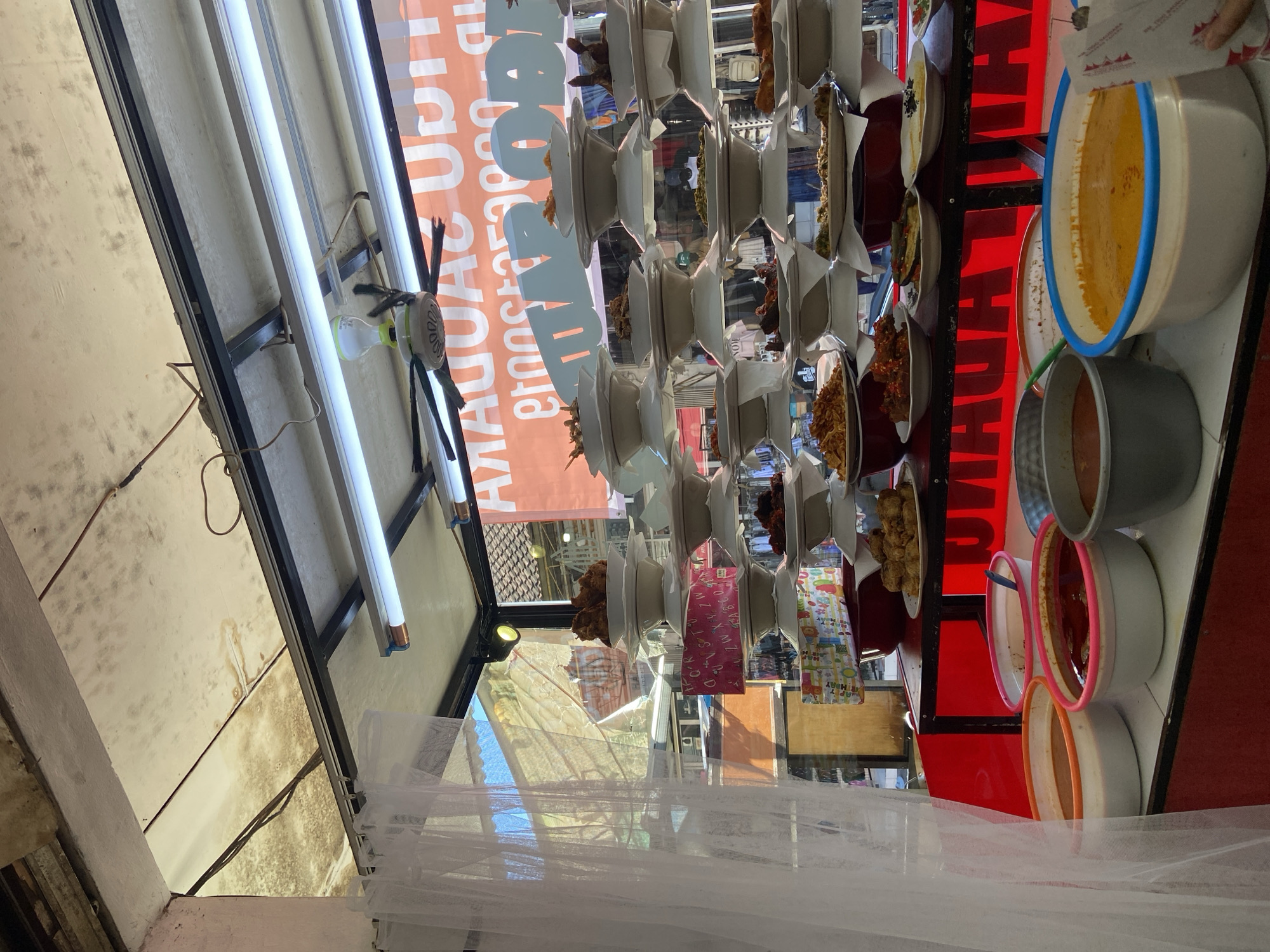 The image captures an outdoor food stall in Kuta, Indonesia. The stall features a variety of Indonesian dishes displayed on shelves. The foreground shows a white, translucent curtain or netting hanging down. Above, there are overhead fluorescent lights and a decorative spider hanging from the light fixture. The stall prominently features an orange sign with white lettering in Indonesian, advertising food offerings. Stacks of white plates hold individual servings of various dishes. Several bowls and pots filled with different sauces and preparations are arranged on the right side of the stall. The background displays signs with text in Indonesian. The overall atmosphere suggests a busy food stall selling local cuisine.