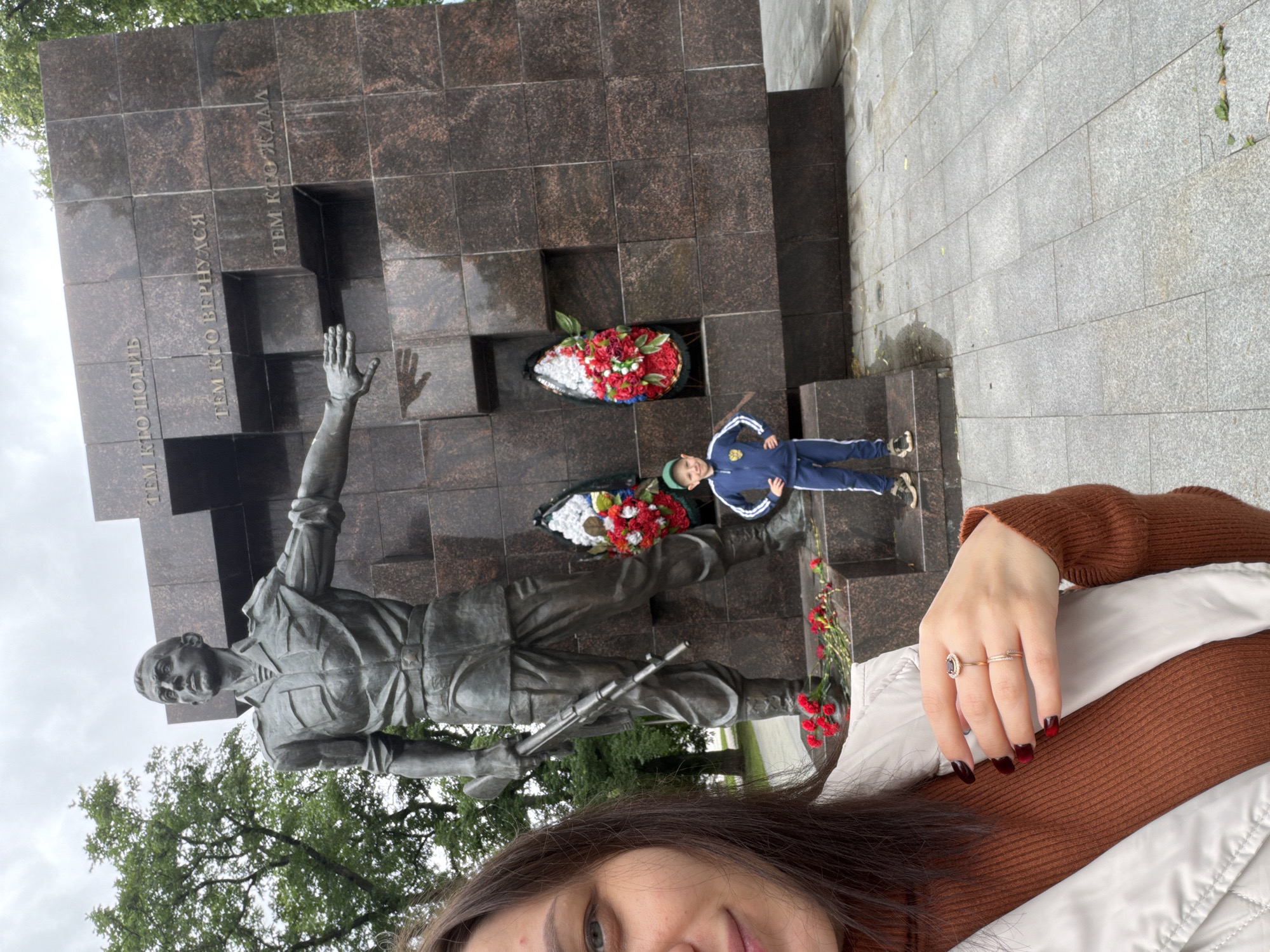 In Perm, Russia, a woman and a young boy stand before a war memorial. The memorial features a large bronze statue of a soldier holding a rifle, with outstretched arm. Behind the statue, a granite monument is inscribed with text. A young boy in a blue tracksuit and a green cap stands to the right of the statue. The woman, in the foreground, is partially in frame, with her hand adorned with rings visible. Red flowers are placed at the base of the memorial. The setting appears to be an outdoor public space with trees and paved ground.