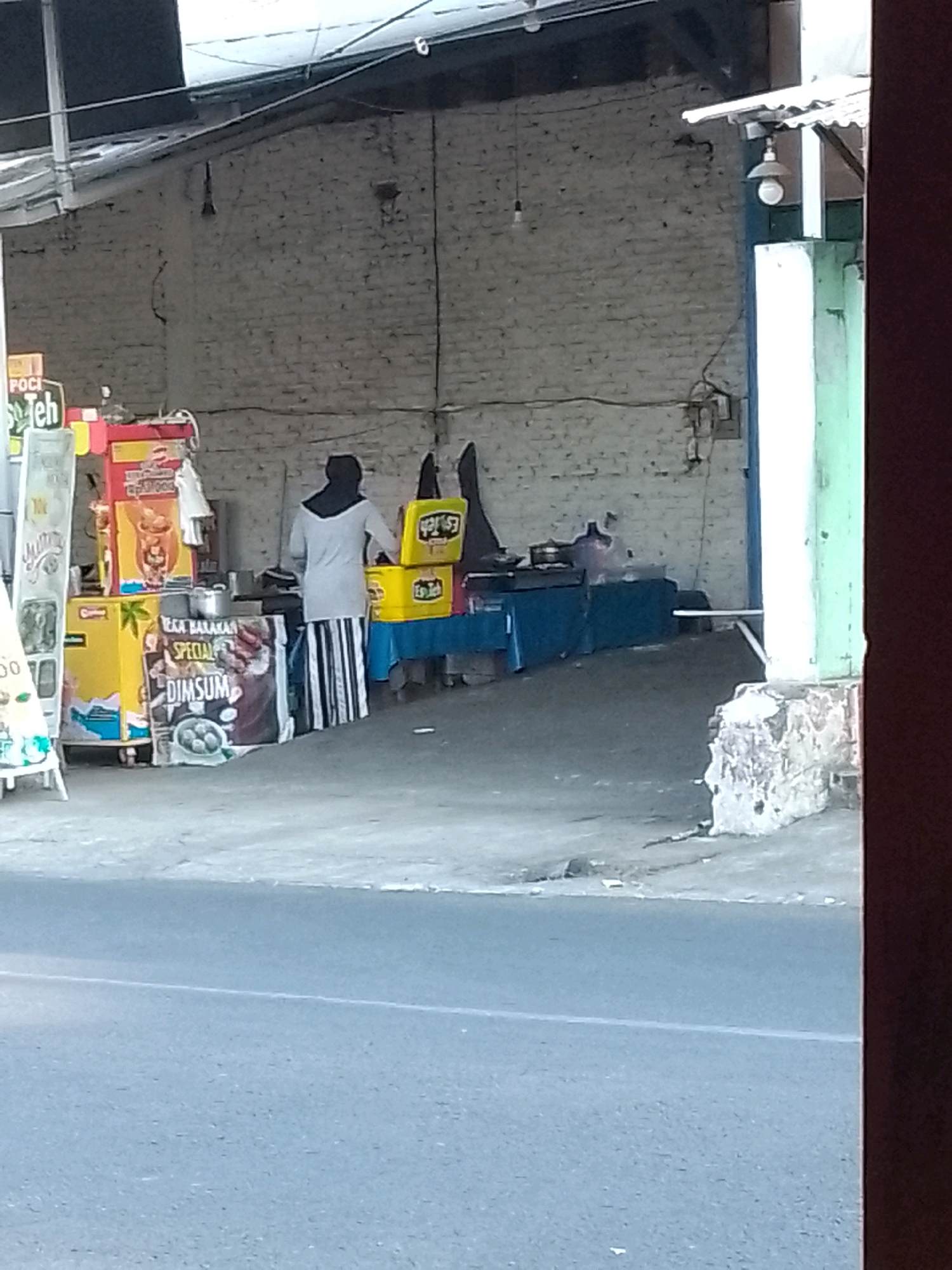 A wide-angle, eye-level shot captures a scene at a roadside stall in Gebang, Indonesia. The stall appears to be selling drinks and possibly food, with various brightly colored containers and signage. A person, dressed in a grey tunic and a black hijab, stands behind the stall, seemingly engaged in serving customers. To the left, a sign displays "POCI Teh," indicating the brand of tea sold. Another sign to the right of the stall reads "Es Teh," suggesting iced tea is also available. A white easel sign with a blurry image is positioned in the foreground on the left. The background consists of a weathered brick wall. The ground is a mix of concrete and asphalt. The image is slightly blurry, suggesting it was taken with a handheld camera.