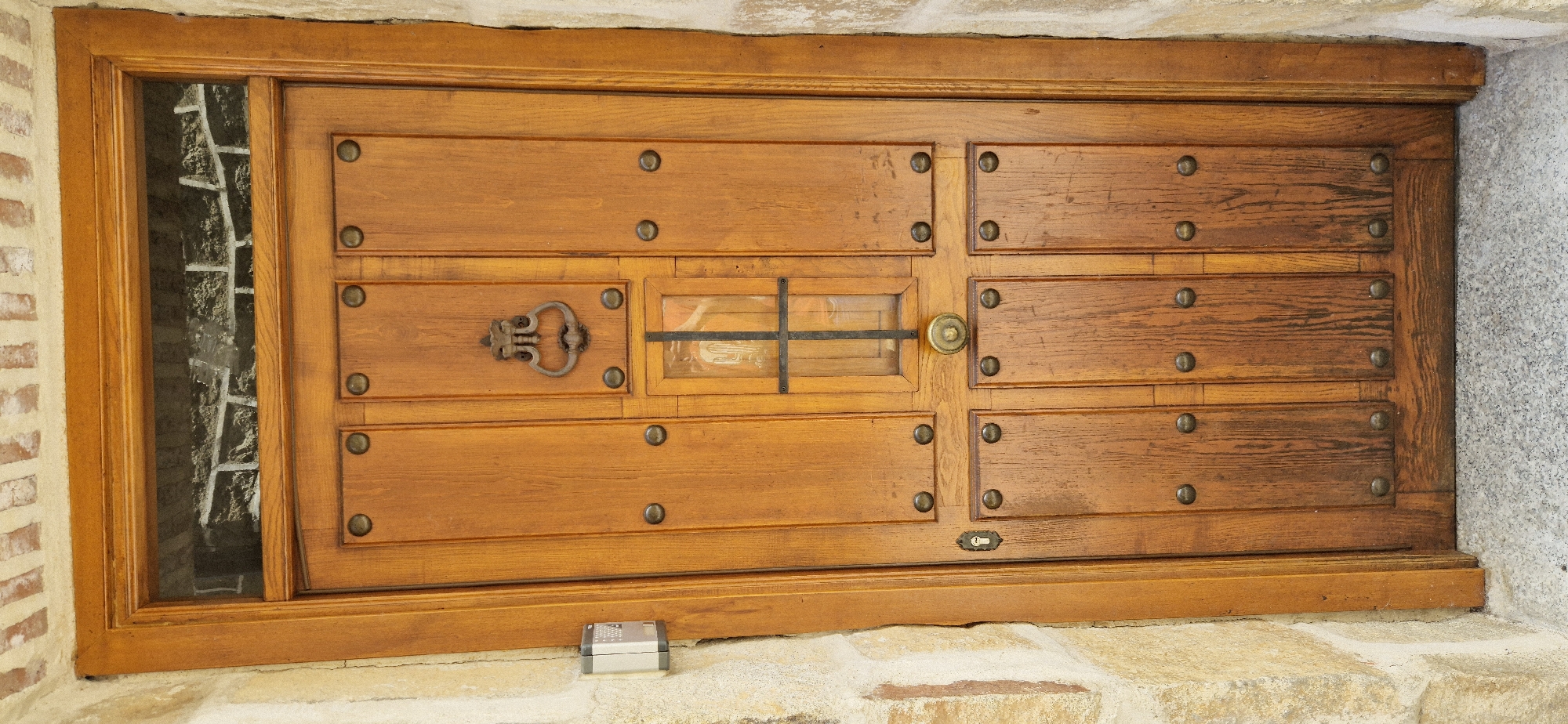 This is a medium shot, eye-level, exterior photograph of a wooden door with a glass insert and metal hardware. The door is set into a stone wall. The door is made of horizontal wooden planks with vertical wooden dividers, and has several round, metallic rivets. There is a decorative iron door knocker on the left side of the door, and a round brass doorknob on the right side. A small, rectangular glass window with a metal crossbar runs horizontally across the center of the door. A small, silver electronic device with a keypad is visible on the stone threshold at the bottom center of the door. The left side of the image shows a narrow, vertical glass pane that allows a view of the brickwork and some metal bars behind it. The lighting suggests it is daytime. The overall impression is of an old, possibly rustic or historic entrance.