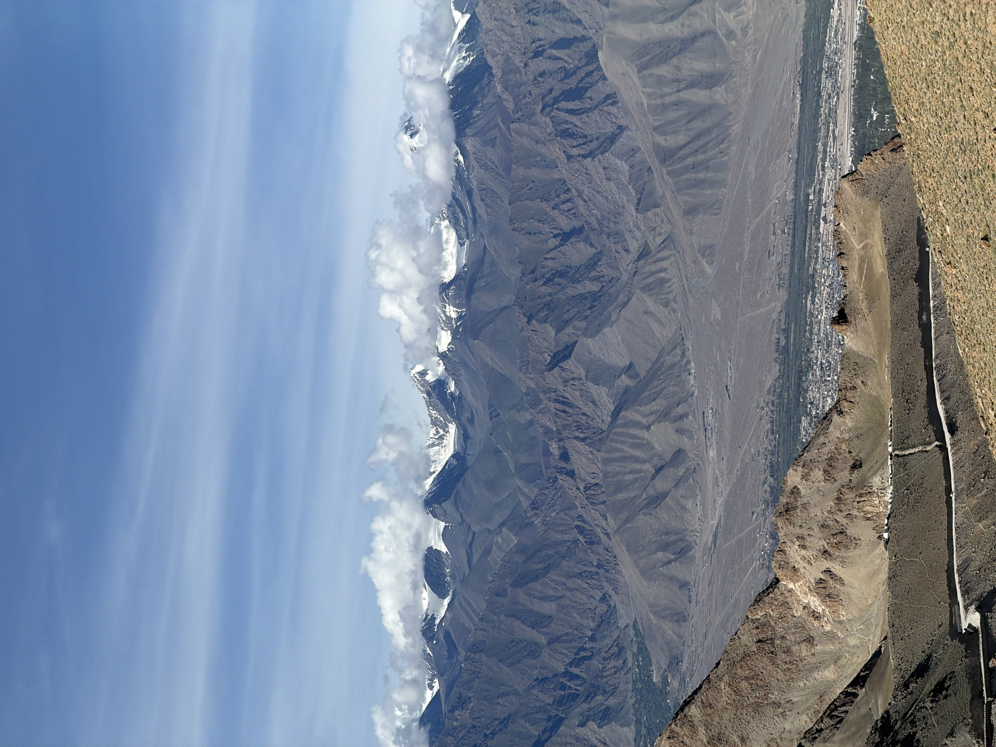 The image captures a panoramic view of Leh, India, with a focus on its mountainous terrain and the town nestled within a valley. The foreground features arid, rocky slopes with sparse vegetation, a winding road, and a few white structures possibly indicative of buildings or shelters. The middle ground reveals the town of Leh, characterized by a dense cluster of buildings with visible rooftops, spread across the valley floor, bordered by patches of green vegetation. Beyond the town, the landscape transitions into vast, rugged mountains, displaying layers of barren, eroded slopes in varying shades of brown and gray. In the background, a majestic range of snow-capped peaks pierces the sky, partially obscured by white clouds. The sky above is a clear blue, with thin, wispy clouds stretching horizontally across the upper portion of the frame. The scene conveys a sense of grandeur and the unique geographical setting of Leh.