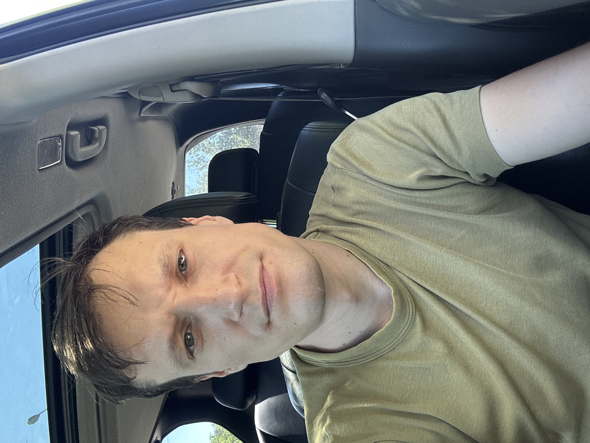 A man is taking a selfie from the driver's seat of a car. He has short brown hair and is wearing a light green t-shirt. The interior of the car is visible behind him, including the headrests of the seats and a sunroof. The background outside the car appears to be a sunny day with trees. The image was taken in the city of Makiv, Ukraine.