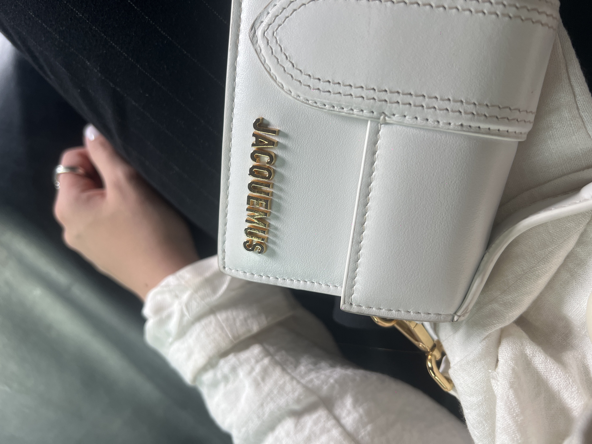 A close-up shot shows a white leather handbag featuring the word "JACQUEMUS" vertically in gold lettering. The handbag has stitched detailing along its edges and gold hardware. A person's hand with a silver ring is partially visible. Parts of a black garment and a white linen shirt or scarf are also visible. The location and city are unknown.