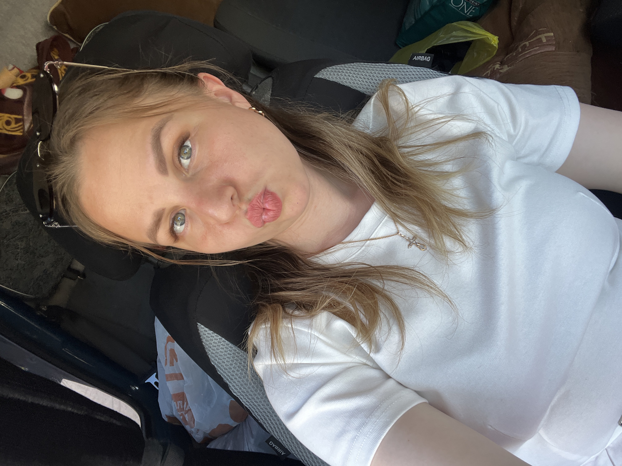 A young woman with light brown hair and green eyes is lying on a black car seat with a grey mesh. She's wearing a white t-shirt. Her lips are pursed. Sunglasses are atop her head. A gold necklace with a cross pendant is visible. An "AIRBAG" tag is visible on the side of the seat. Part of a brown pillow and green pet food bag are in the background. Part of a white plastic bag is by the car door. The location is not discernible from the image and cannot be identified as Izhevsk, Russia.