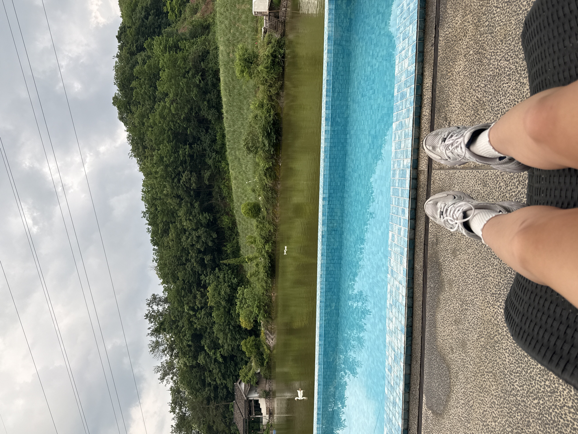 A vertical, eye-level shot depicts a landscape next to a pool, with the photographer’s legs and silver sneakers appearing in the foreground, situated on what looks like a patterned outdoor seating area.
Beside the pool is a murky body of water, possibly a lake or river, with a reflection of the surrounding trees and sky visible on its surface. There is also a small structure, possibly a boathouse, seen on the bank. Two white waterfowl float in the lake.
Across the body of water, a dense forest rises up a hillside, with power lines visible in the cloudy sky above. Based on the prompt, the location is identified as being in the city of Jinhua, China.