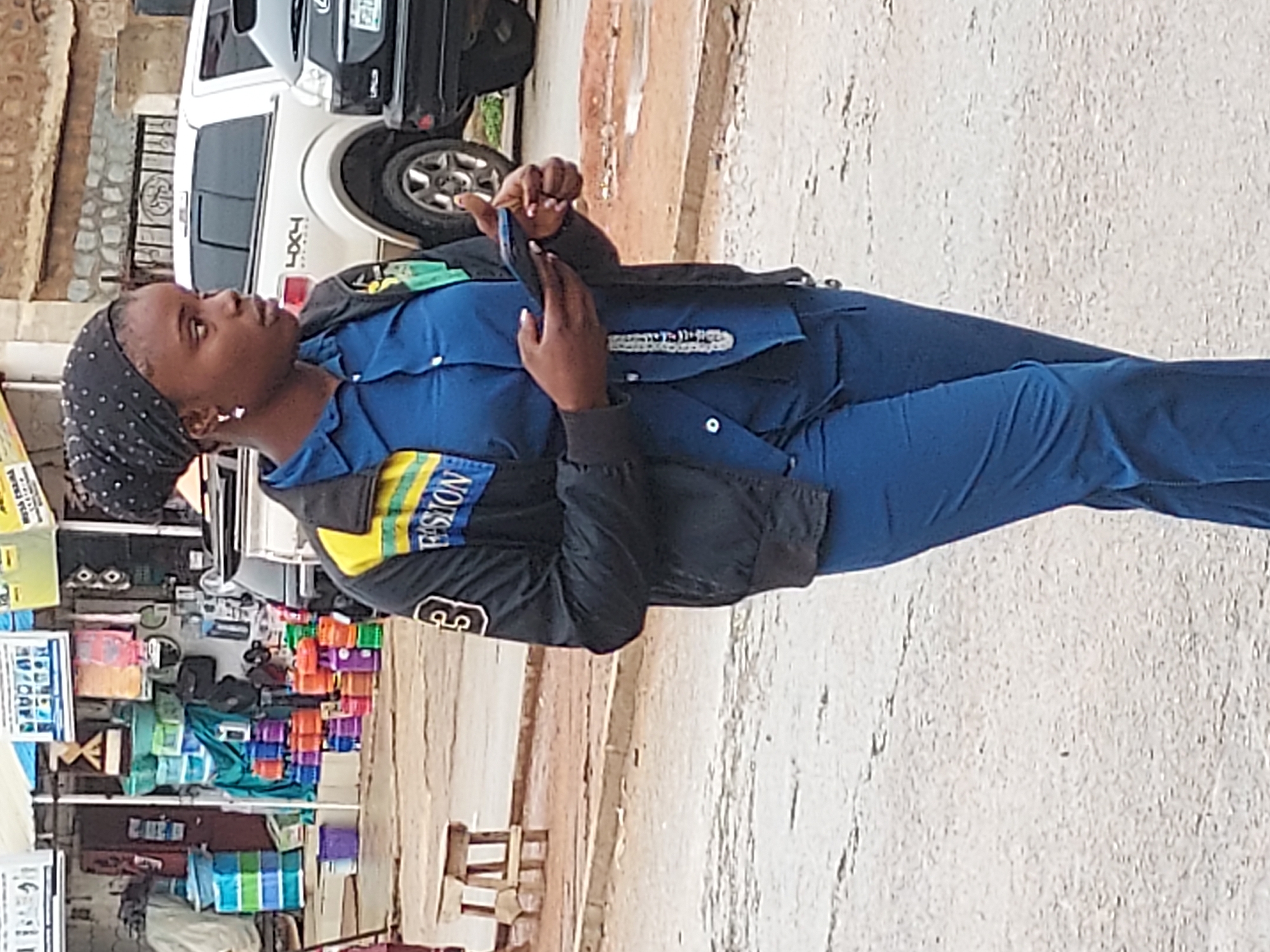 A vertical, full-body outdoor shot captures a young woman standing on what appears to be a street or roadside in Jos, Nigeria, while looking at her smartphone. She is positioned slightly to the left of the frame, facing right, with her head tilted upwards as if looking down at the screen. Her expression is neutral and focused.
She wears a dark blue uniform-style top with matching trousers, layered with a black jacket. The jacket features a yellow and light green stripe on the left sleeve with partial text "FISION" (likely "FUSION") and a white numeral "3" or "E" below it. A dark, polka-dotted headscarf covers her hair. Her hands, adorned with light-colored nail polish, hold the smartphone.
In the background, a white SUV with "FX4" emblazoned on its side is parked, partially obscured by a dark-colored car behind it. A stone-patterned wall is visible behind the vehicles. To the lower left, a vibrant street vendor's stall or shop displays numerous colorful items, possibly plastic containers or packaged goods, stacked neatly. A wooden stand or table is also visible near the stall. The ground is a light-colored, possibly paved or dusty surface. The scene is brightly lit, suggesting daytime. Signs near the stall contain blurry text, with "WORLD OF..." and "WAZIRI" vaguely discernible on one, and a possible "BAAZAL BASARU" on another.