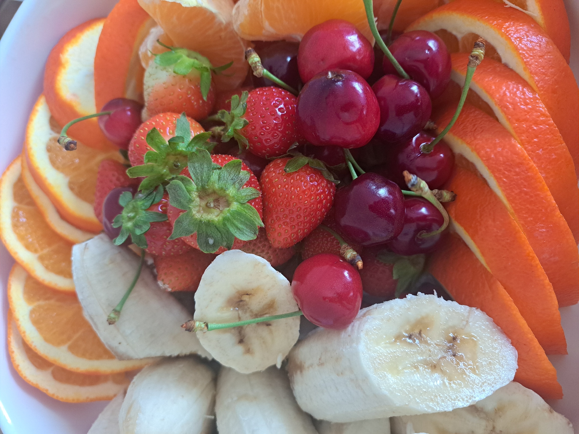 This close-up image displays an assortment of fresh, prepared fruits arranged within a white, shallow container. The scene is located in the city of Artik, Armenia. The arrangement includes numerous segments of peeled oranges, presenting as semicircular slices with bright orange pulp and a thin rind, visible along the top and bottom perimeters. Interspersed on the left side are multiple peeled banana pieces, cut into thick, circular or oval cross-sections, exhibiting a pale yellow to off-white color with minor brownish discoloration at the center of some slices, indicative of oxidation. The central and right-central areas contain a cluster of whole, ripe strawberries, characterized by their bright red color and intact green calyxes. Among the strawberries and extending to the lower-right are several dark red to deep maroon cherries, each retaining a slender green stem. The fruits are densely packed, filling the frame. No individuals are present, and no specific actions are in progress. The lighting is even, illuminating the natural textures and colors of the produce.
