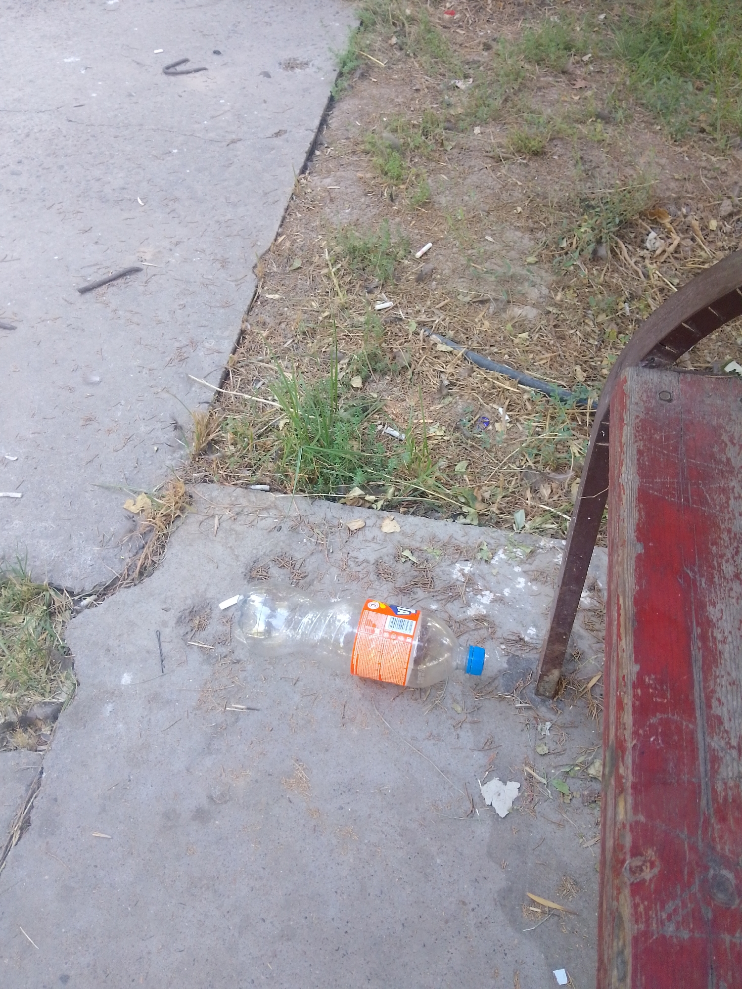 The image depicts an outdoor scene in Абай ауылдық округі, Kazakhstan, characterized by a concrete walkway adjacent to a patch of dry grass and dirt. A red wooden bench, weathered and showing signs of age, is positioned on the right side of the frame. On the concrete, a plastic bottle with a blue cap and an orange label is lying on its side. Scattered debris, including what appears to be cigarette butts and small pieces of paper, are visible on the ground. The overall impression is one of a neglected public space.