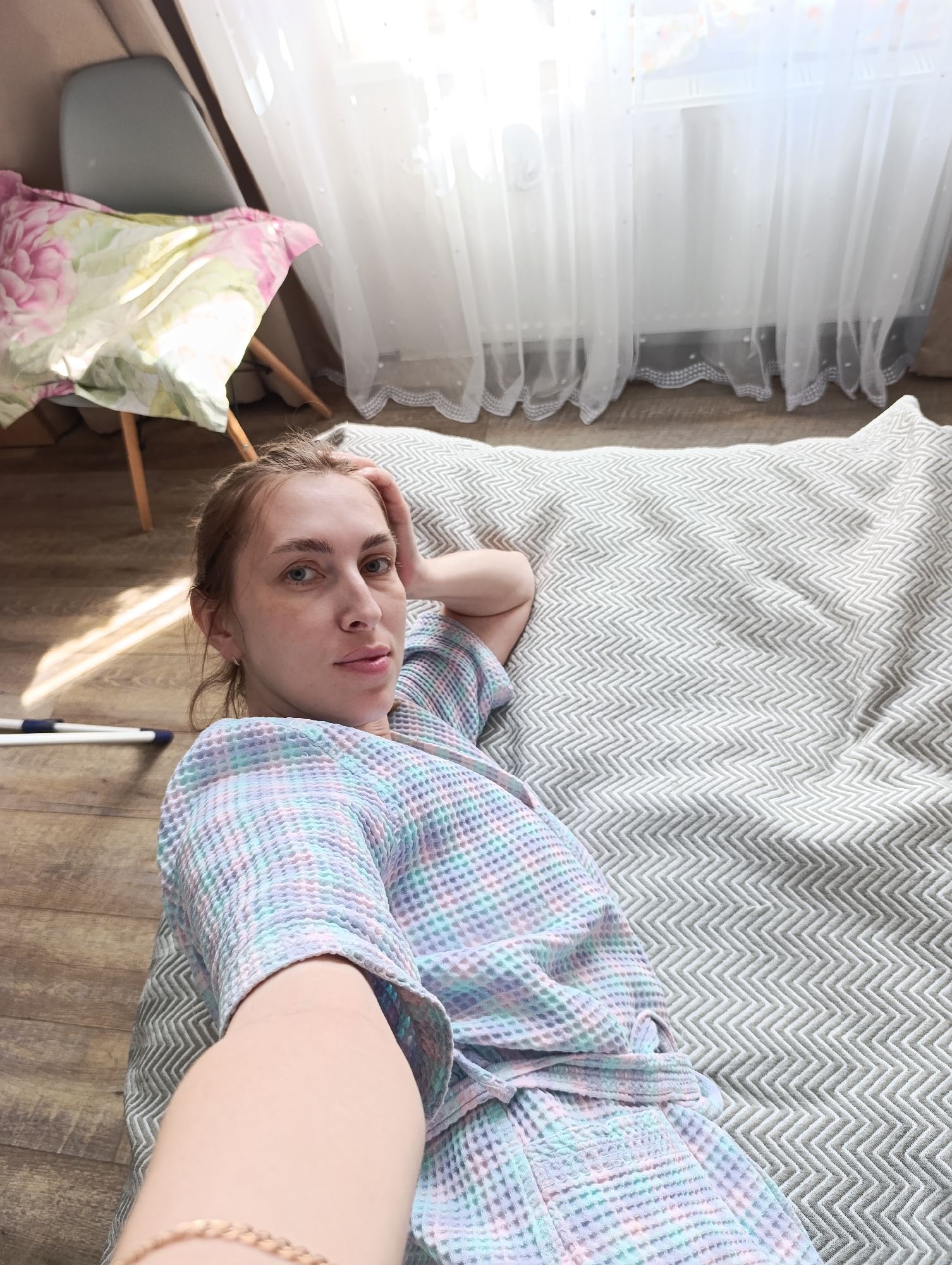 A woman is lying on a patterned gray blanket on a wooden floor in a room. She is wearing a multicolored pastel robe. Her right arm is bent and her hand is touching the top of her head. Her left arm is extended towards the camera. The location is Chaikovsky, Russia. A chair with a floral cushion sits behind her and a window with white curtains is to her right. A cleaning tool is on the floor to her left.