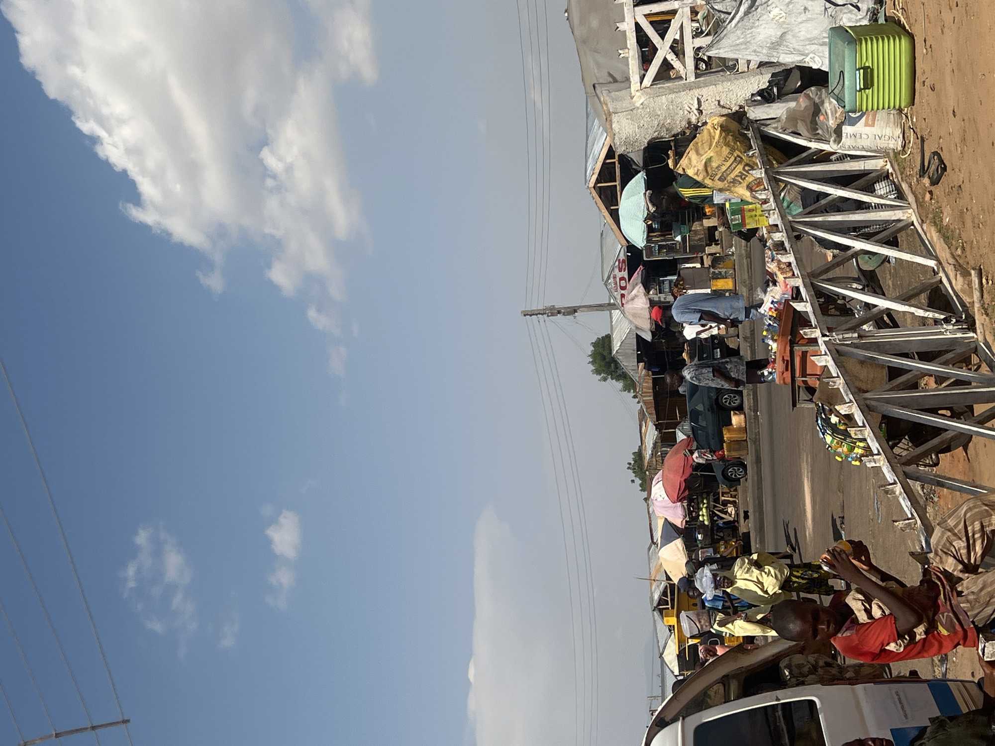 The image depicts an active outdoor market scene in Gombe, Nigeria, under a bright blue sky with scattered white cumulus clouds. Multiple overhead power lines span horizontally across the upper portion of the frame. The foreground consists of uneven, reddish-brown dirt ground, transitioning to a paved asphalt road in the mid-ground.
Numerous market stalls, constructed with corrugated metal roofs and various wooden supports, line the street. Several colorful market umbrellas, including red, light blue, and beige, provide shade over various vendor areas. A white van or bus, with its side door open, is partially visible on the far left. A dark-colored sedan is parked on the road in the mid-right, beyond the market stalls.
A white metal barrier or fence, composed of horizontal bars and vertical supports, extends across the lower-mid foreground. Approximately ten individuals are visible throughout the scene. In the immediate foreground, a young male wearing a red t-shirt and striped trousers is seated on the white metal barrier, holding a clear plastic bottle containing an orange liquid. To his left, another individual in a dark green shirt is partially visible.
Behind the white barrier, a large yellow sack, partially labeled "BETA HARVEST," rests on the ground. A bright green plastic cooler box is situated on the dirt ground in the lower-right, adjacent to a white sack, partially labeled "NGAI CEME." A black sandal or slipper lies on the ground nearby.
In the mid-ground, several individuals are present, standing or walking. One person near the white van wears a yellow garment and has a white plastic container on their head. Further into the market, a person in a blue shirt stands near a stall shaded by a red umbrella. Another individual is visible near the parked dark car. A pile of watermelons or similar fruits is displayed on a distant market stall.