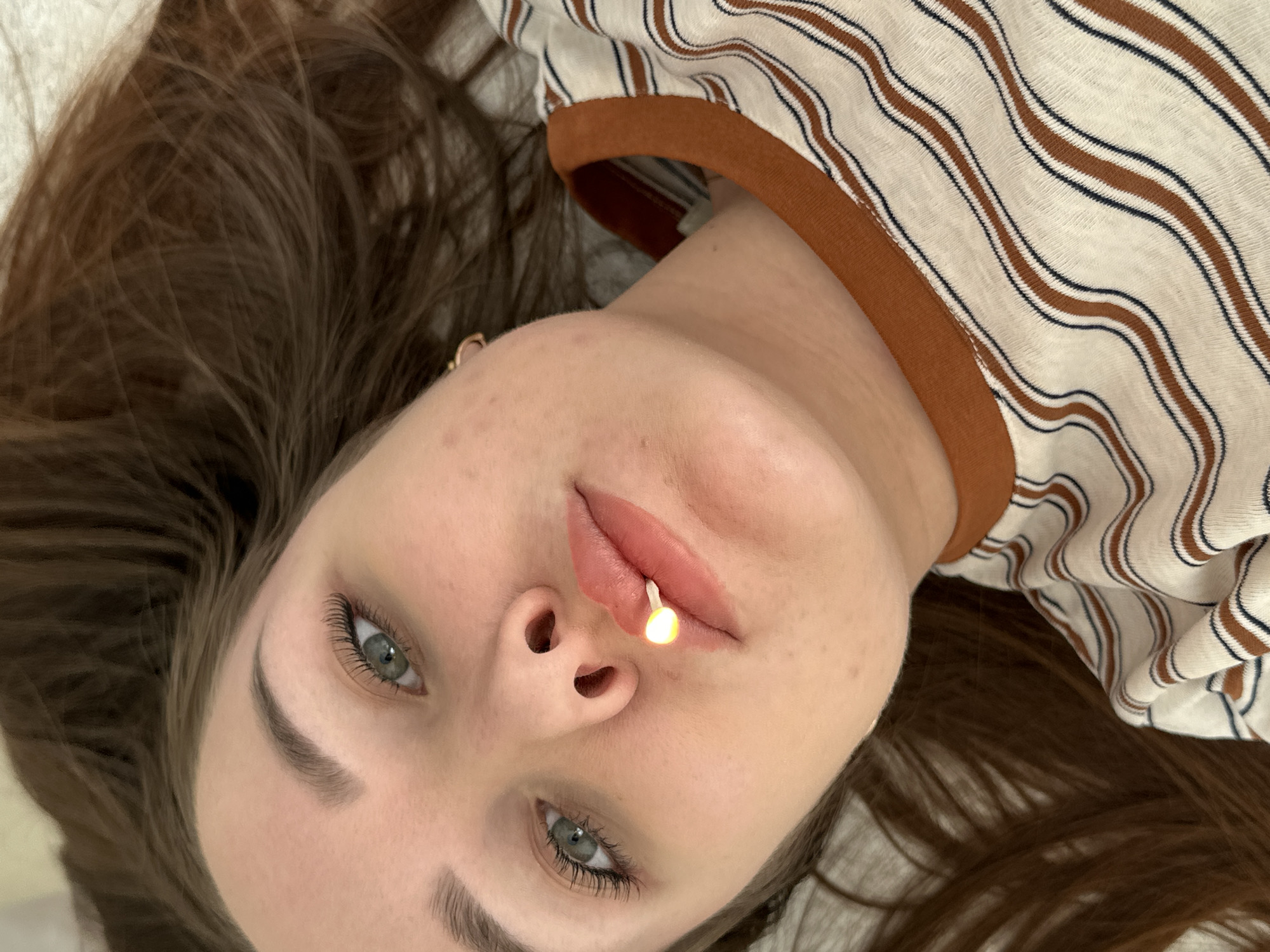 A young woman is lying down, with her head tilted towards the viewer. She has long brown hair and fair skin, with a lighted sparkler held between her lips. She is wearing a striped shirt with a brown neckline. The background is plain and out of focus. The location, Chaikovsky, Russia, cannot be confirmed from the image alone.