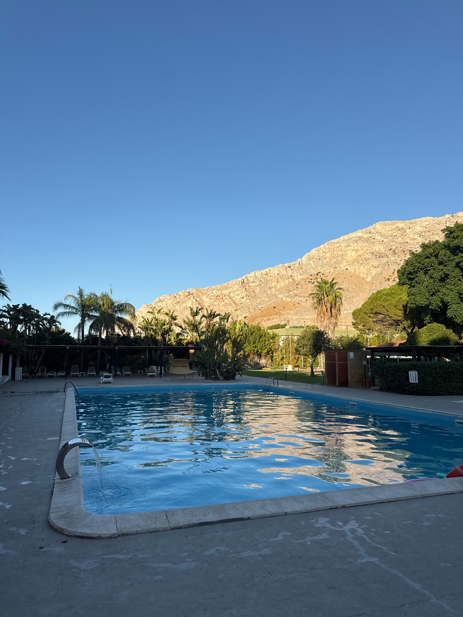 The image captures a serene outdoor swimming pool area at what appears to be a resort or hotel in Isola delle Femmine, Italy. The pool itself is filled with clear blue water and has a contemporary chrome waterfall feature flowing into it from the left side. The pool is surrounded by a concrete deck, which shows some light staining. In the background, beyond the pool and deck, is a lush green garden area with various palm trees and other tropical foliage. Further back, a large, rugged, sun-drenched mountain range dominates the horizon, its textured surface illuminated by natural light. The sky above is a clear, vibrant blue, indicating fair weather. There are no visible people actively using the pool or immediate area, though a few lounge chairs are visible in the shaded garden section. The overall scene suggests a tranquil setting, likely in the late afternoon or early morning due to the angle of the sunlight on the mountains.