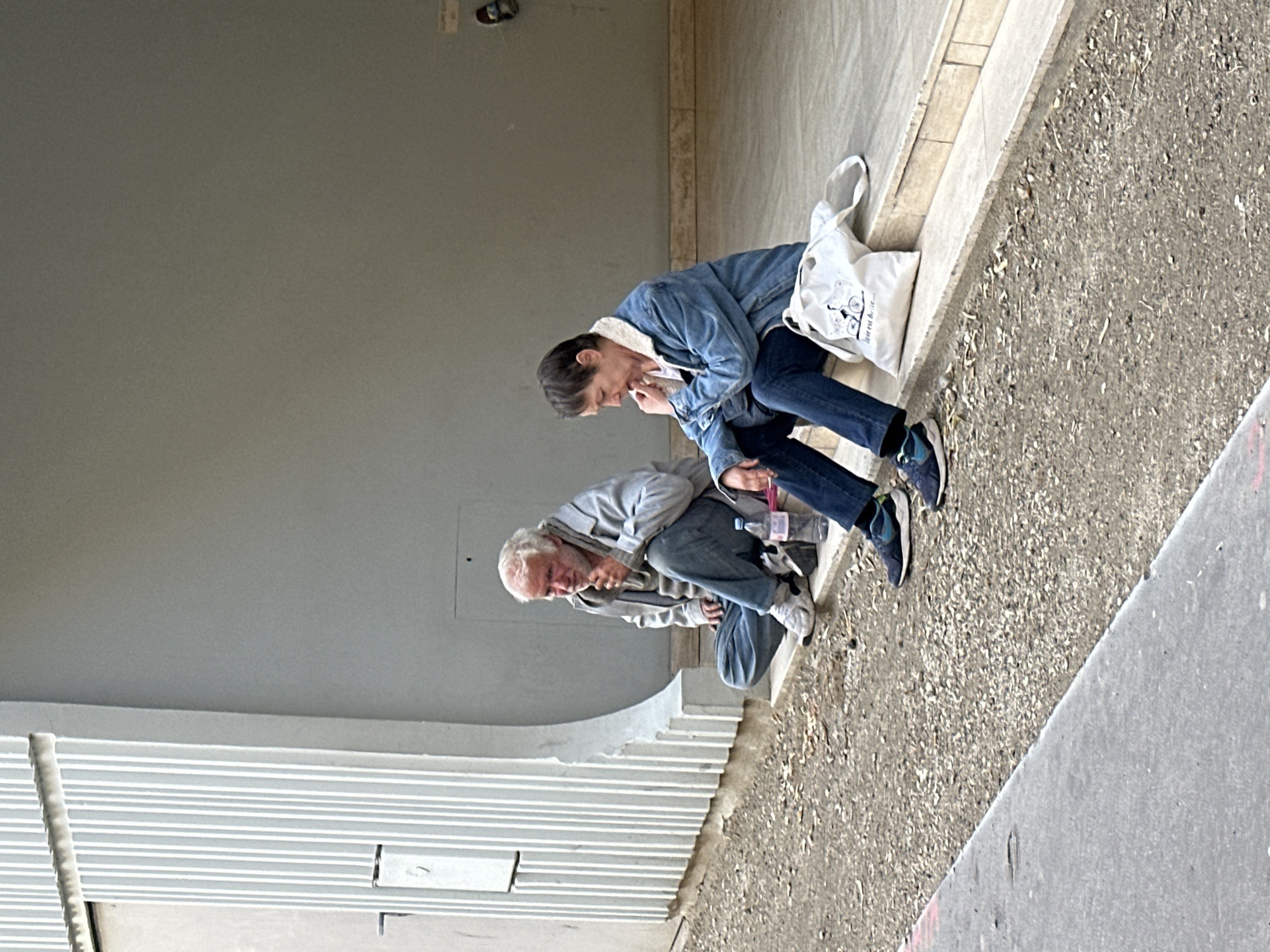 Two people are seated near a sidewalk. One individual, in a denim jacket with a white collar, sits next to a white shopping bag. The other person wears a grey jacket. They both seem to be either resting or engaged in some sort of personal activity. The background features a grey wall and a textured ground surface. The location appears to be an urban setting, possibly in Paris, France.