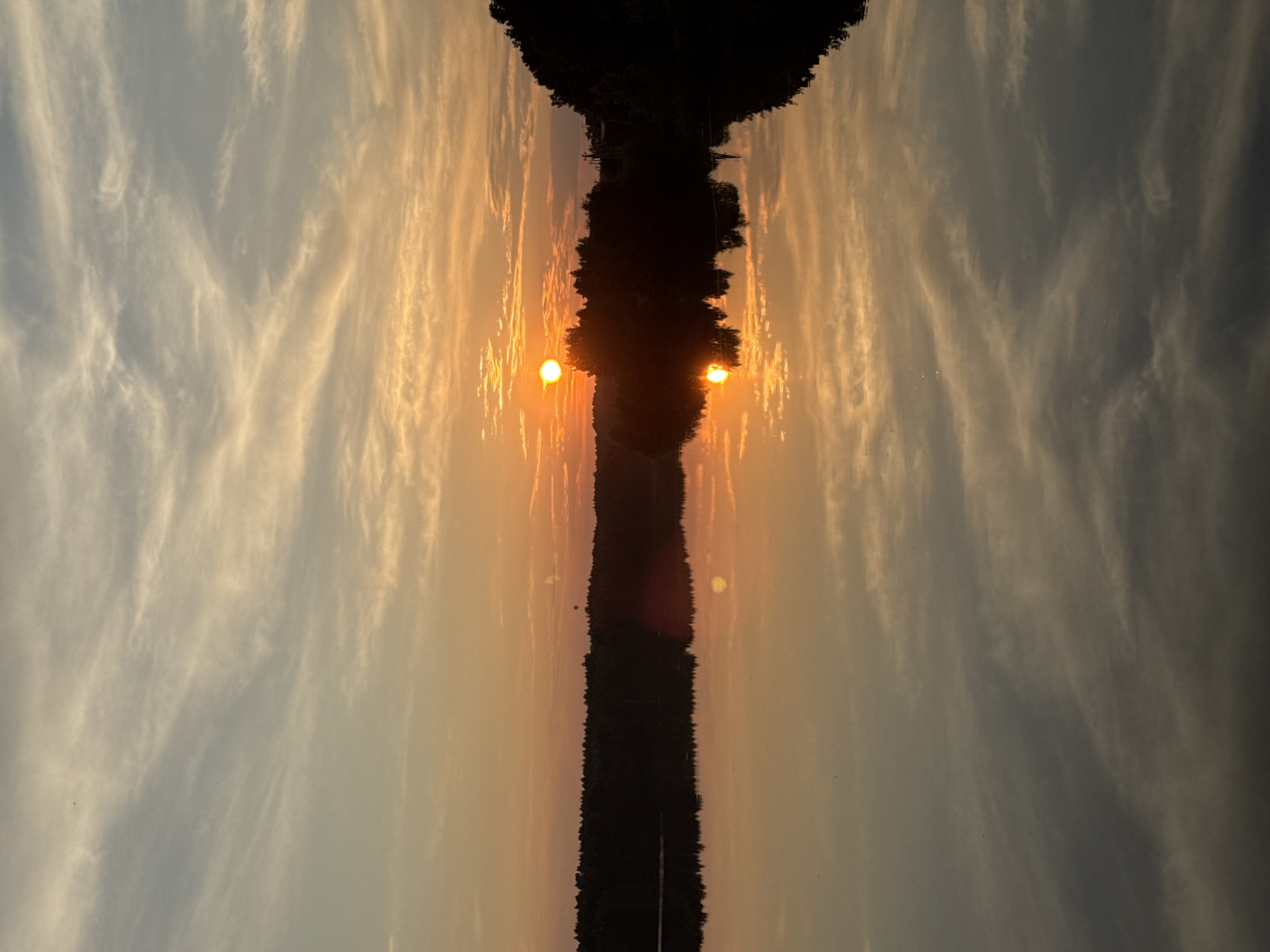 The image depicts a serene sunset over a calm body of water, likely a lake or river, in Kamysh, Russia. The sky is painted with warm hues of orange and yellow, streaked with wispy clouds that reflect beautifully on the water's surface. A silhouetted treeline borders the water, with a few faint structures visible in the distance. The sun, a bright orb, is partially obscured by trees, casting a radiant glow and a distinct reflection on the water. There are no people or animals visible in the scene, and no apparent actions or interactions taking place. The environment is tranquil and natural, characterized by the interplay of light, water, and vegetation.