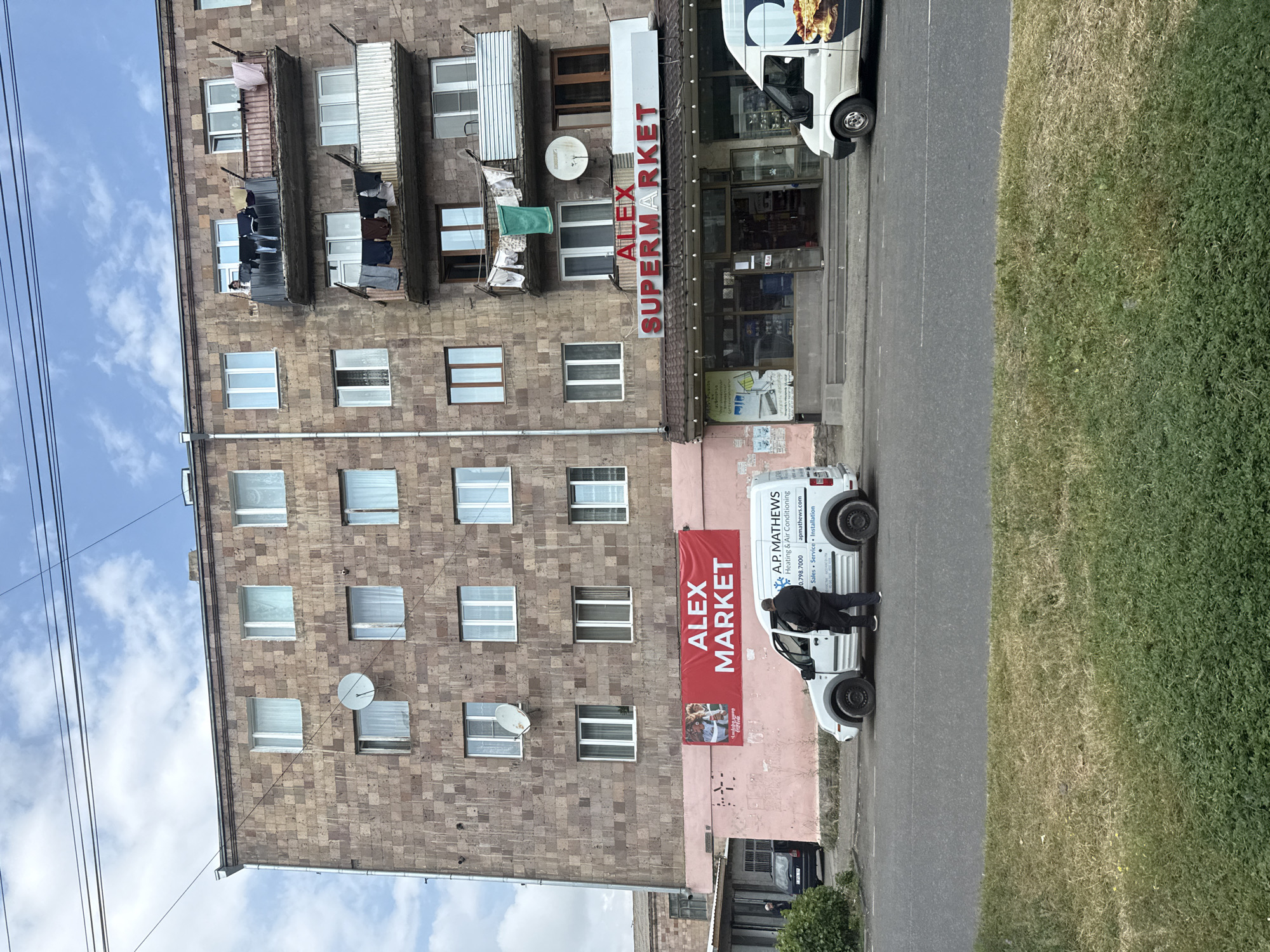 This is a full-frame, eye-level, outdoor, daytime, medium-shot photograph of a building facade in Sevan, Armenia. The building is multi-story, constructed with pinkish-brown stone blocks. There are several windows visible on each floor, some with white curtains. Satellite dishes are mounted on the exterior walls. Balconies on the upper floors have laundry hanging on clotheslines.
On the ground floor, a storefront is visible with the signage "ALEX SUPERMARKET" in red lettering. The storefront has large glass windows displaying merchandise. Two vans are parked in front of the building on the street. One is a white van with the logo "A.P. MATHEWS Heating & Air Conditioning" and a website address. A person is standing next to this van, near the open driver's side door. The second van, partially visible on the left, is dark-colored. A large red banner with "ALEX MARKET" is stretched across the lower part of the building, above the supermarket entrance. A bus stop sign is visible next to the red banner. The foreground features a grassy median strip. The sky is partly cloudy.